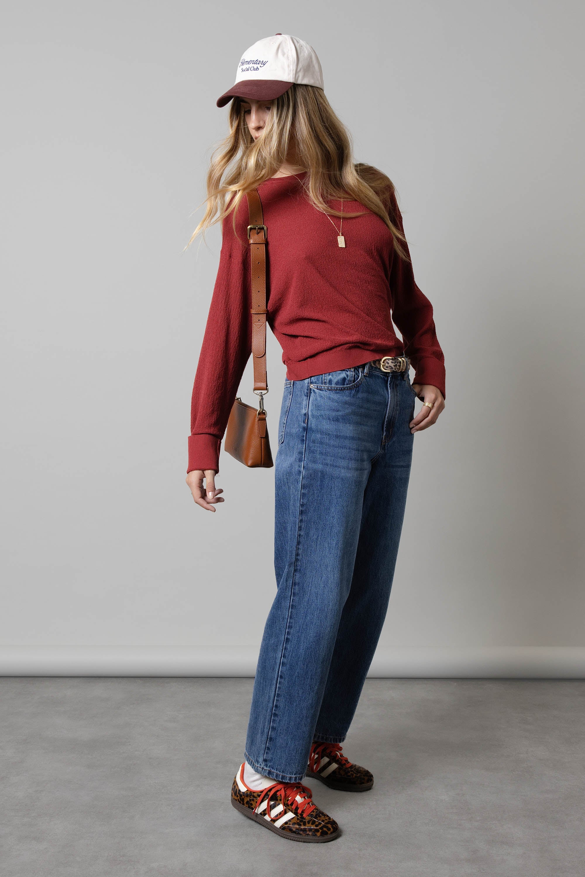 Woman in red top, Max Borrowed From The Boys Jeans, red sneakers, and white cap by gray wall.