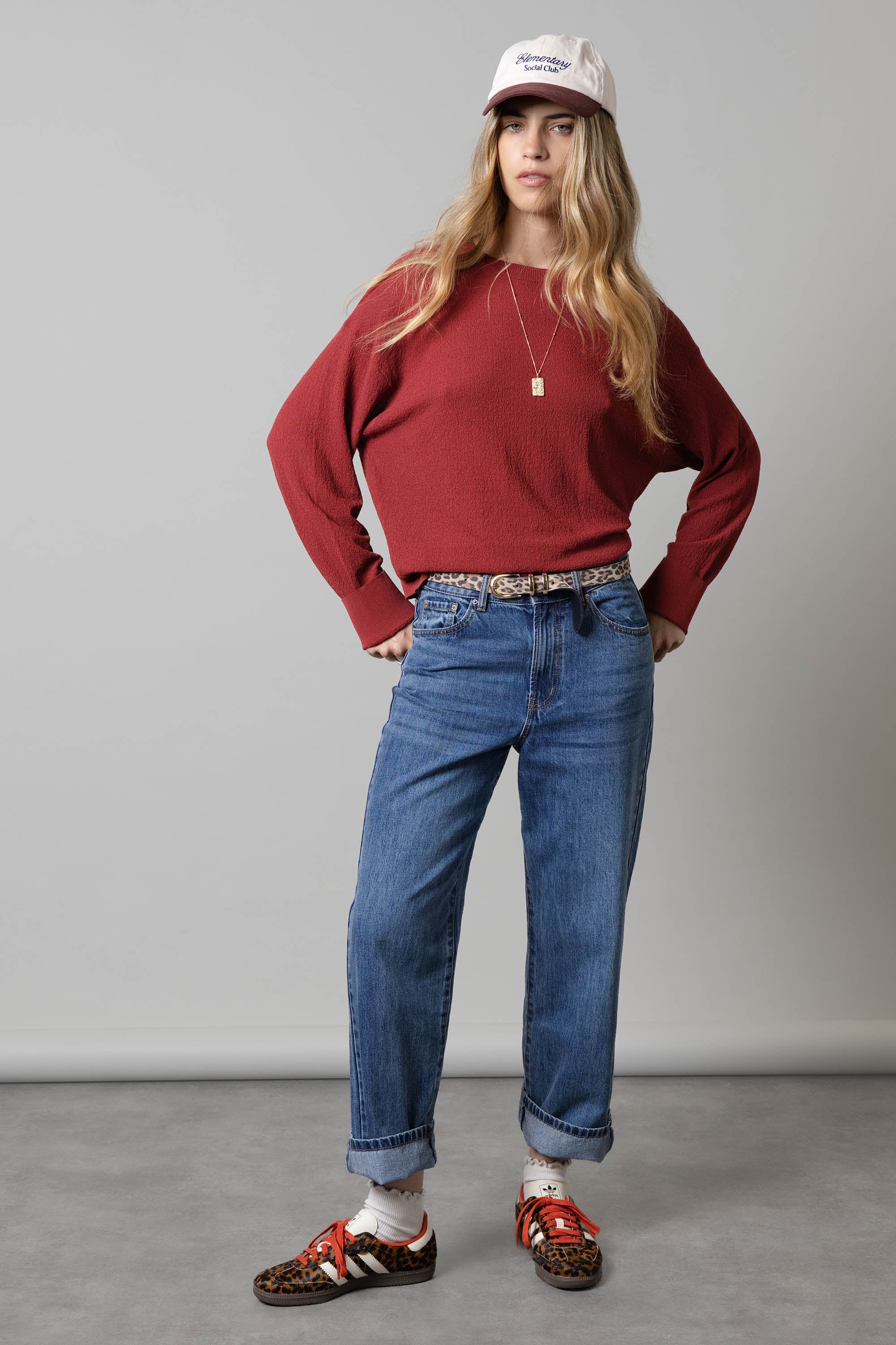 Woman in red sweater, Max Borrowed From The Boys Jeans, sneakers, white cap, and necklace indoors.