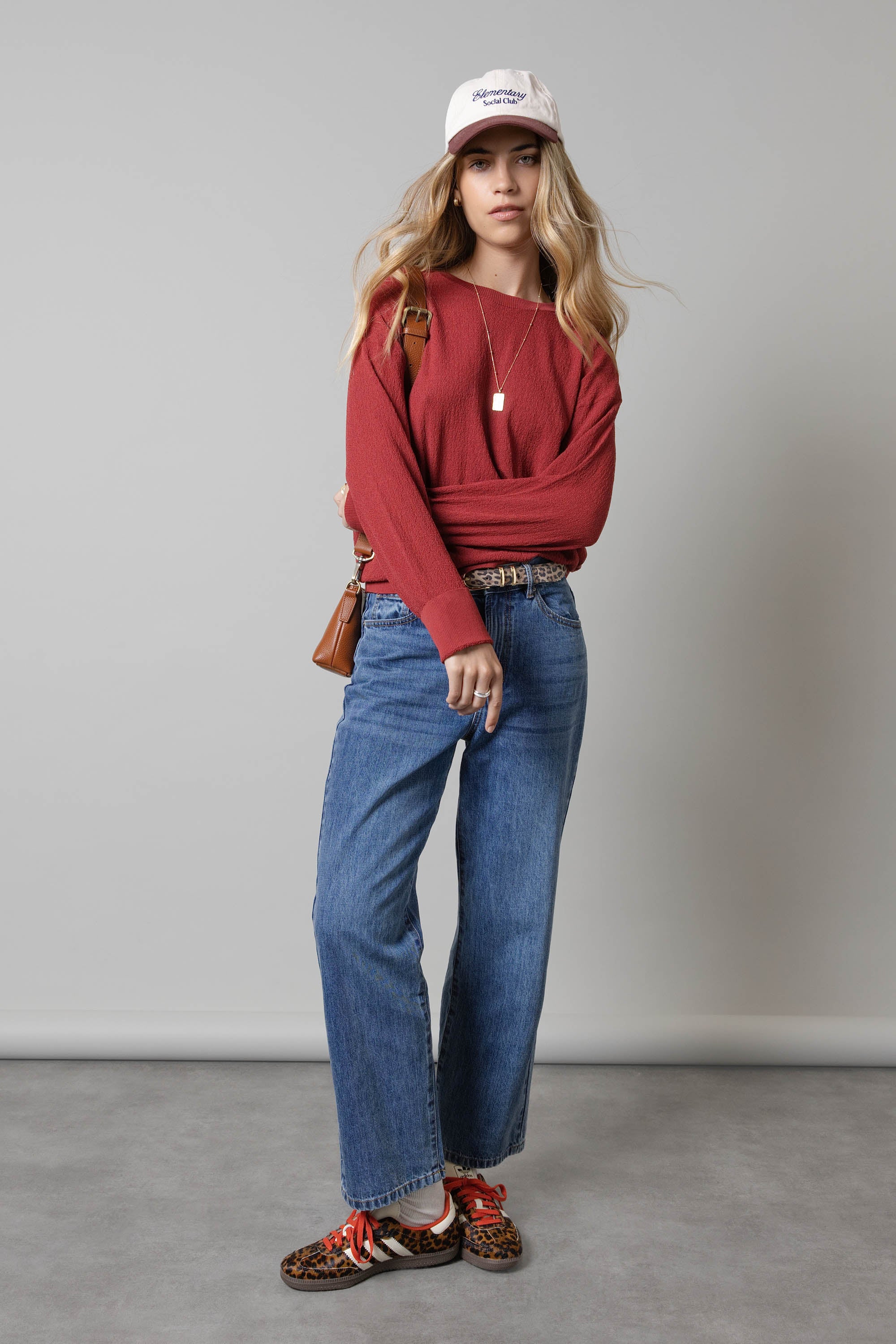 Woman wears a red sweater, white cap, Max Borrowed From The Boys Jeans, and leopard shoes indoors.