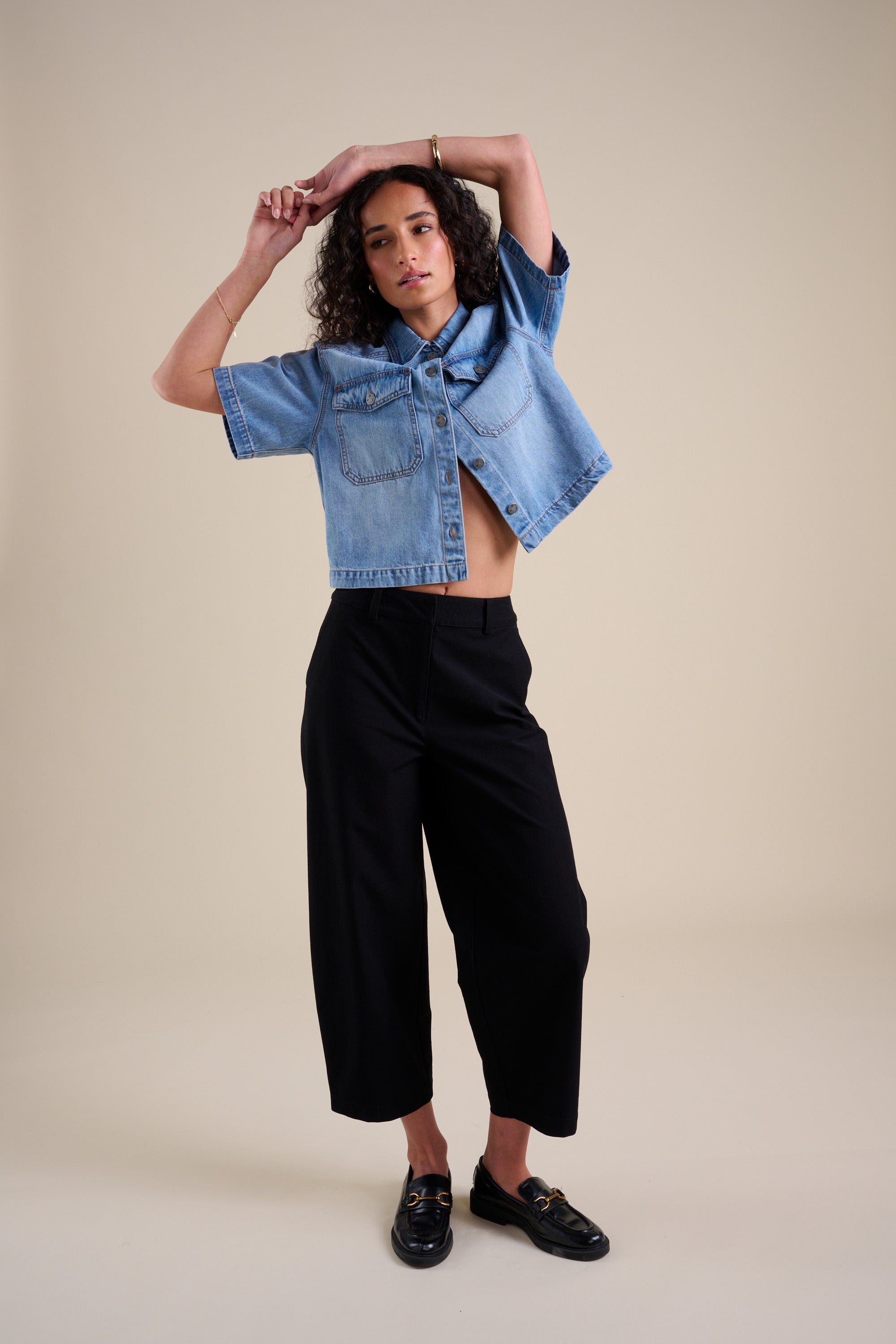 Woman in Eden Barrel Leg Pants by Max poses against a beige background in a denim short-sleeve jacket.