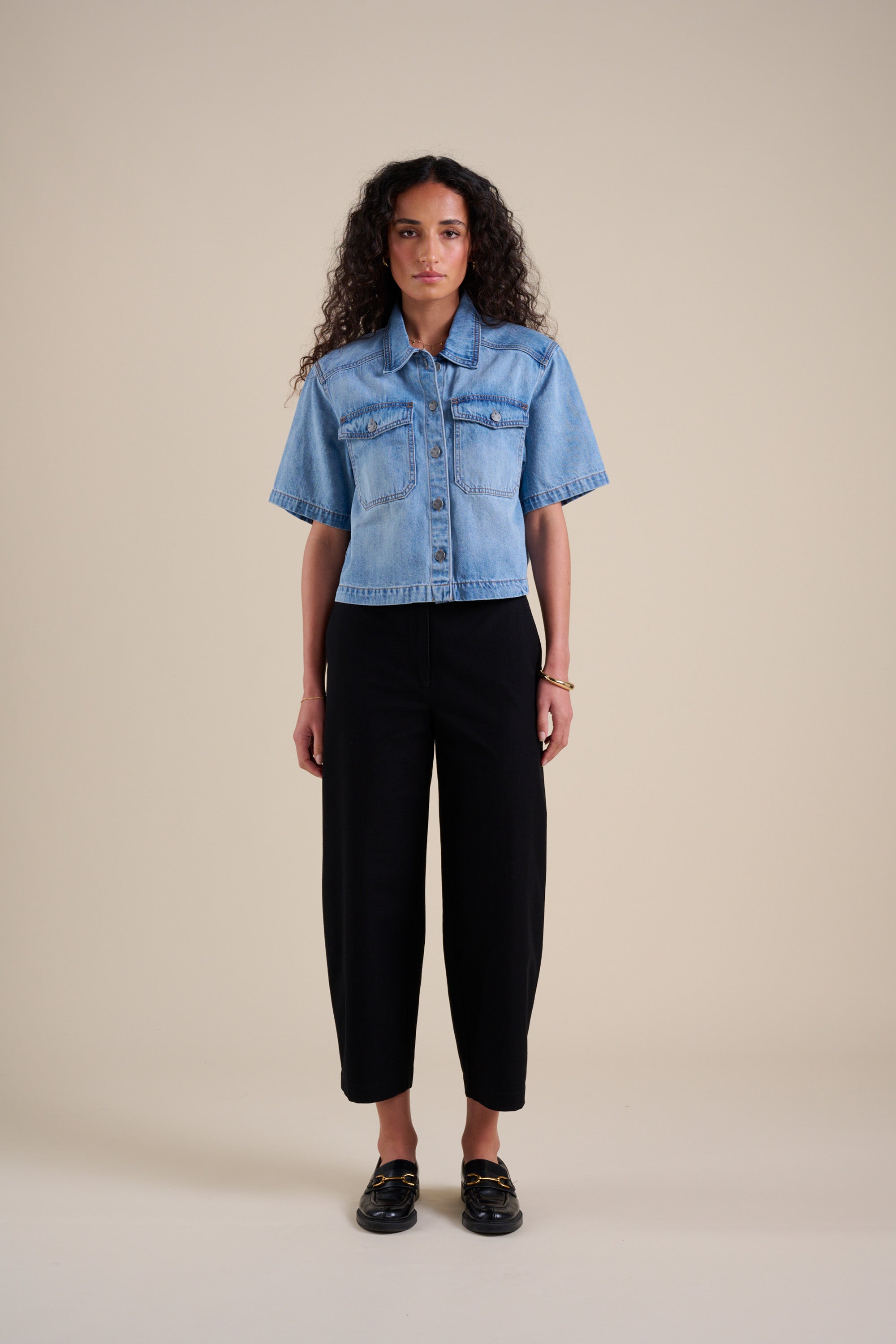 Woman in Eden Barrel Leg Pants by Max and a short-sleeve denim shirt, standing against beige.