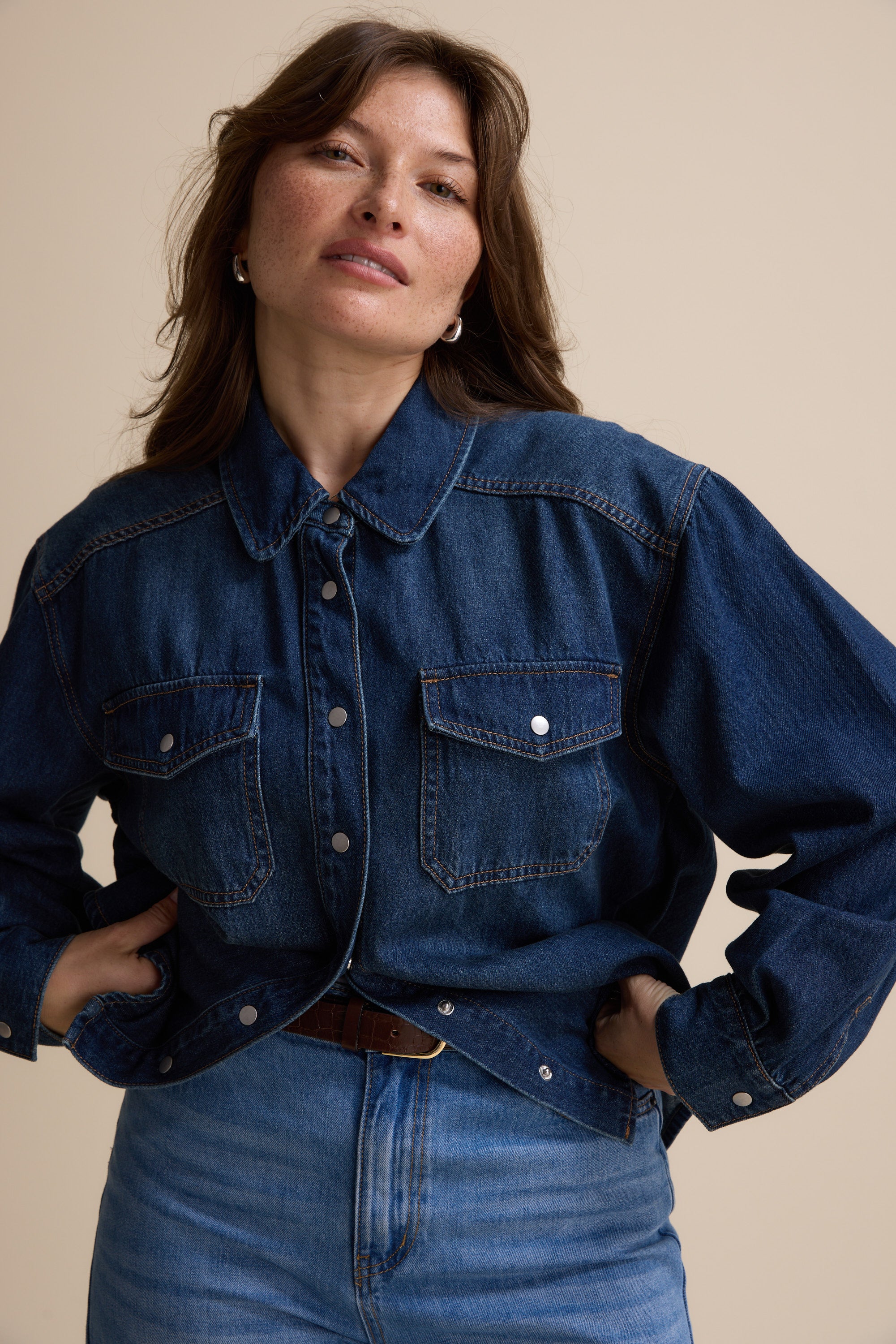 Model poses in Max's Sabrina Cropped Denim Jacket, indigo wash, against a beige background.