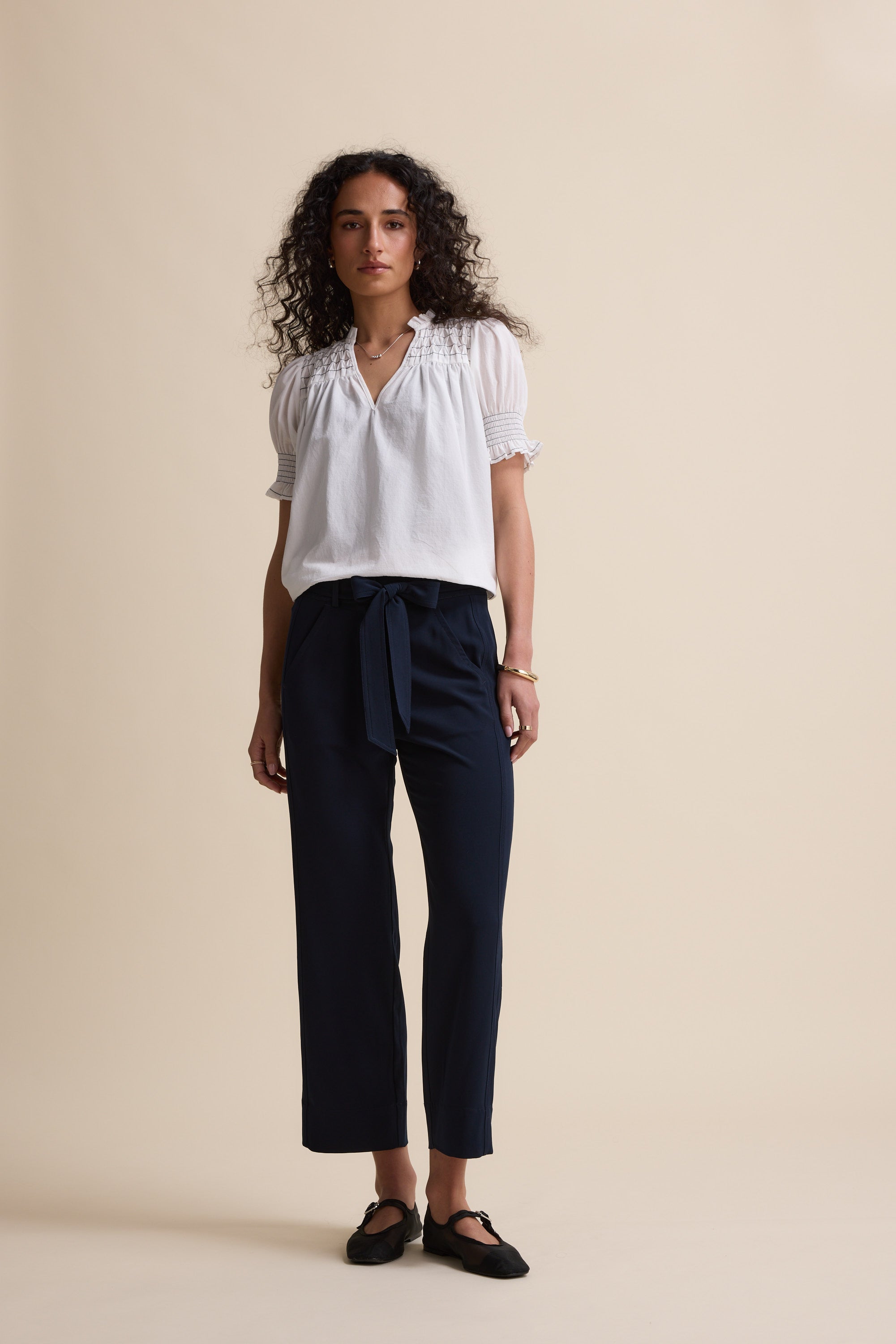 Woman in white blouse and Max's The Easy Care Belted Pants stands against a beige background.