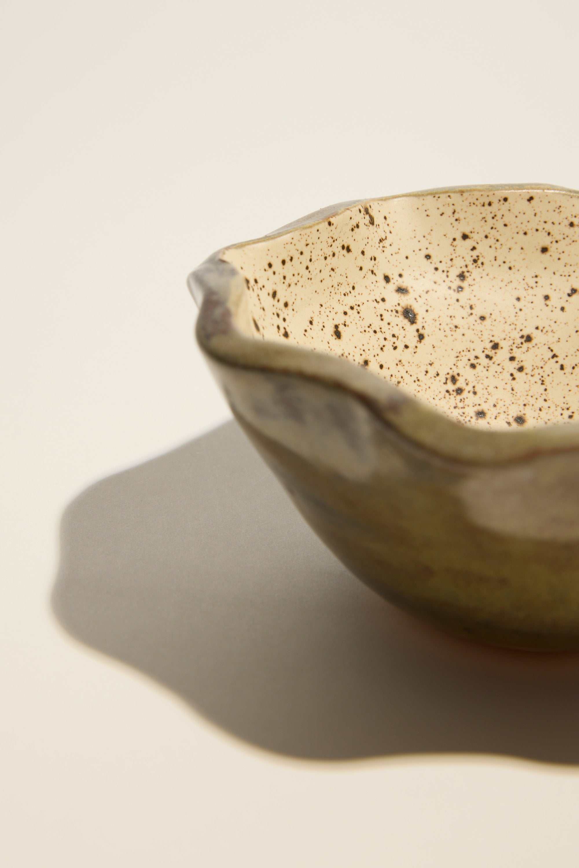 A close-up of the Magnolia Lane Ruffled Condiment Bowl by Magnolia Lane, handmade in NZ with a wavy rim and speckled cream interior, casting a gentle shadow on a light surface.