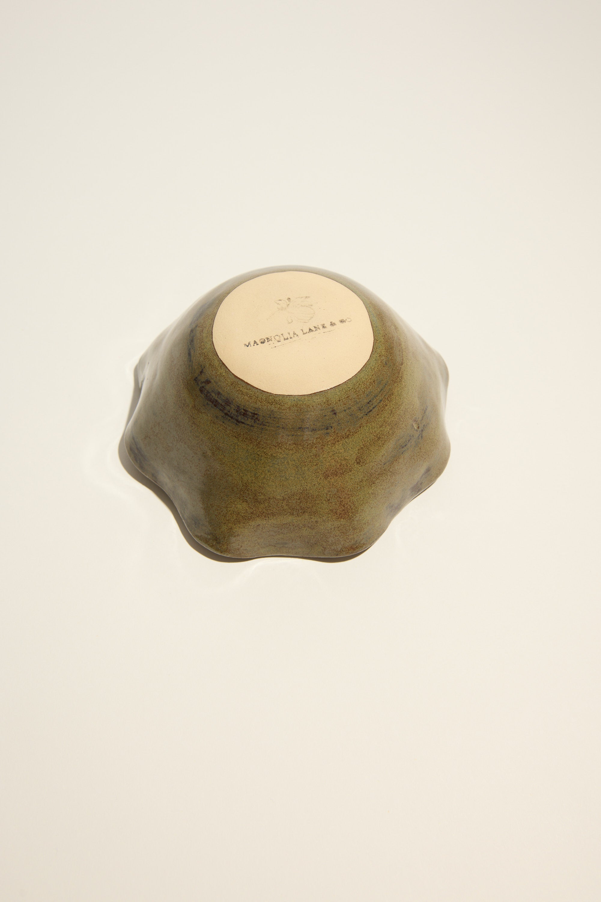 A small Magnolia Lane Ruffled Condiment Bowl sits upside down on a plain, light surface. Its unglazed base shows the Magnolia Lane maker’s mark—artisanal tableware handmade in New Zealand.