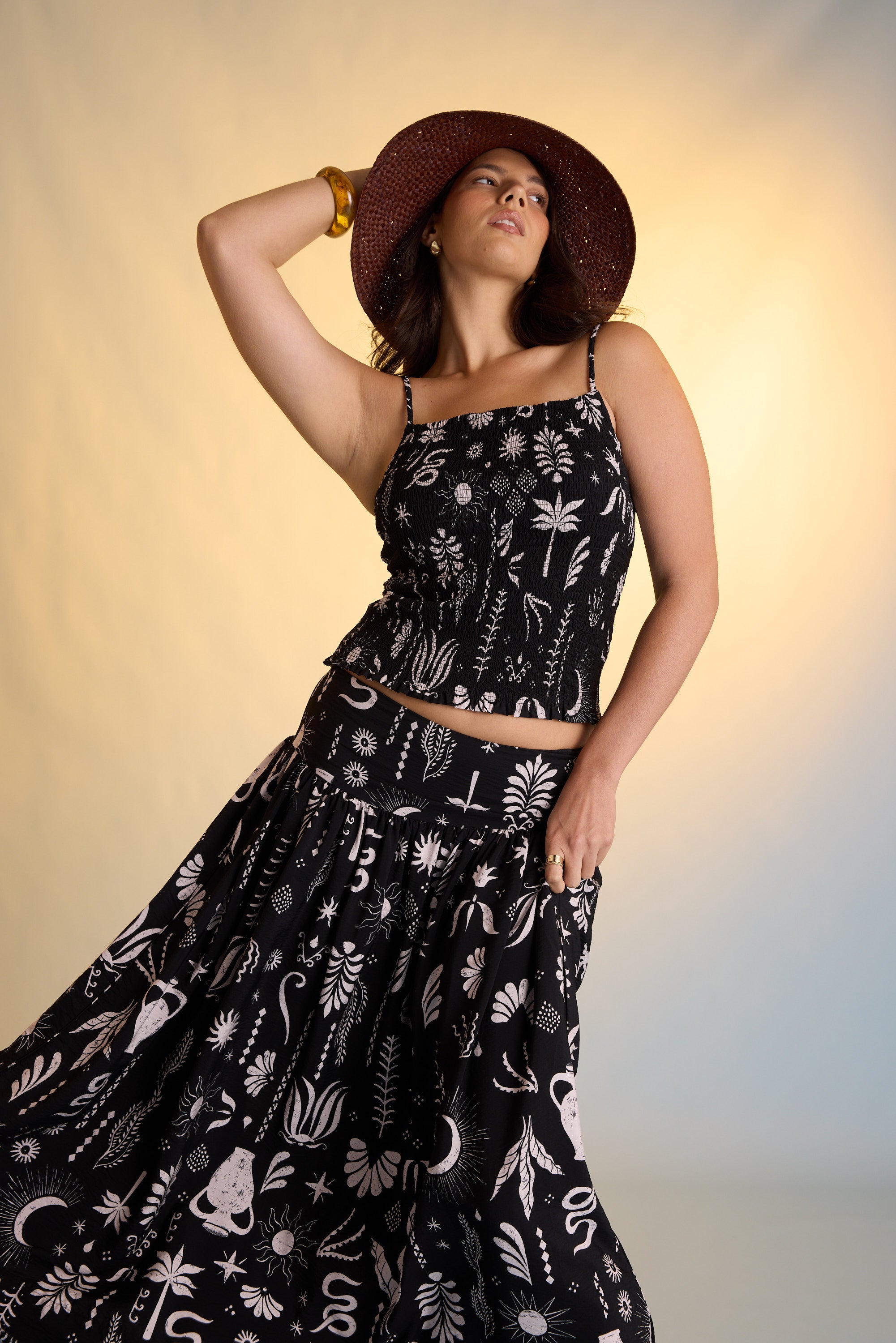 A woman poses confidently in the softly lit background, wearing Max’s Helena Drop Waist Skirt with a black sleeveless top and wide-brimmed hat; the skirt features an exclusive white botanical and animal print.