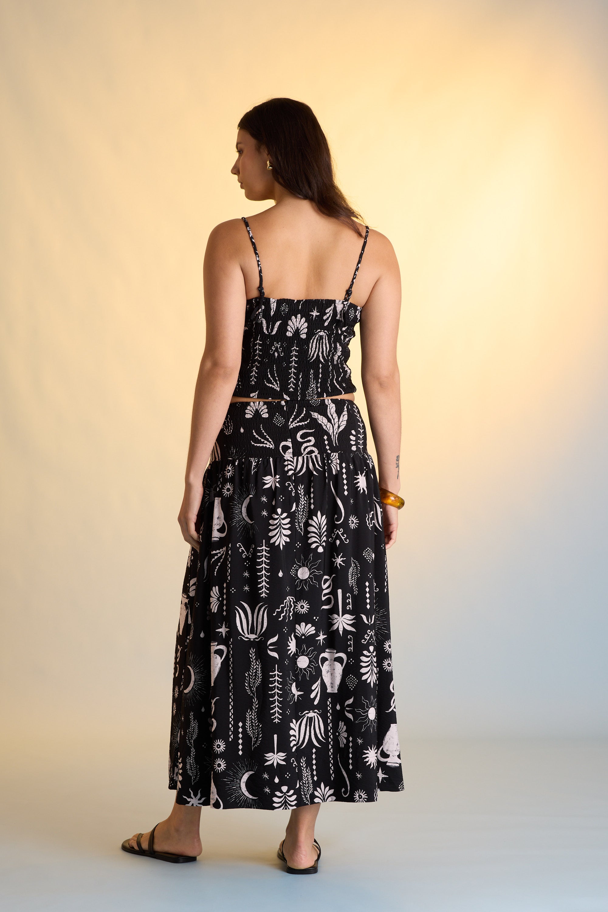 A woman with long dark hair wears the Max Helena Drop Waist Skirt, featuring an exclusive white abstract and botanical print, paired with a black sleeveless top, black sandals, and a brown bracelet. The softly lit background enhances the look.