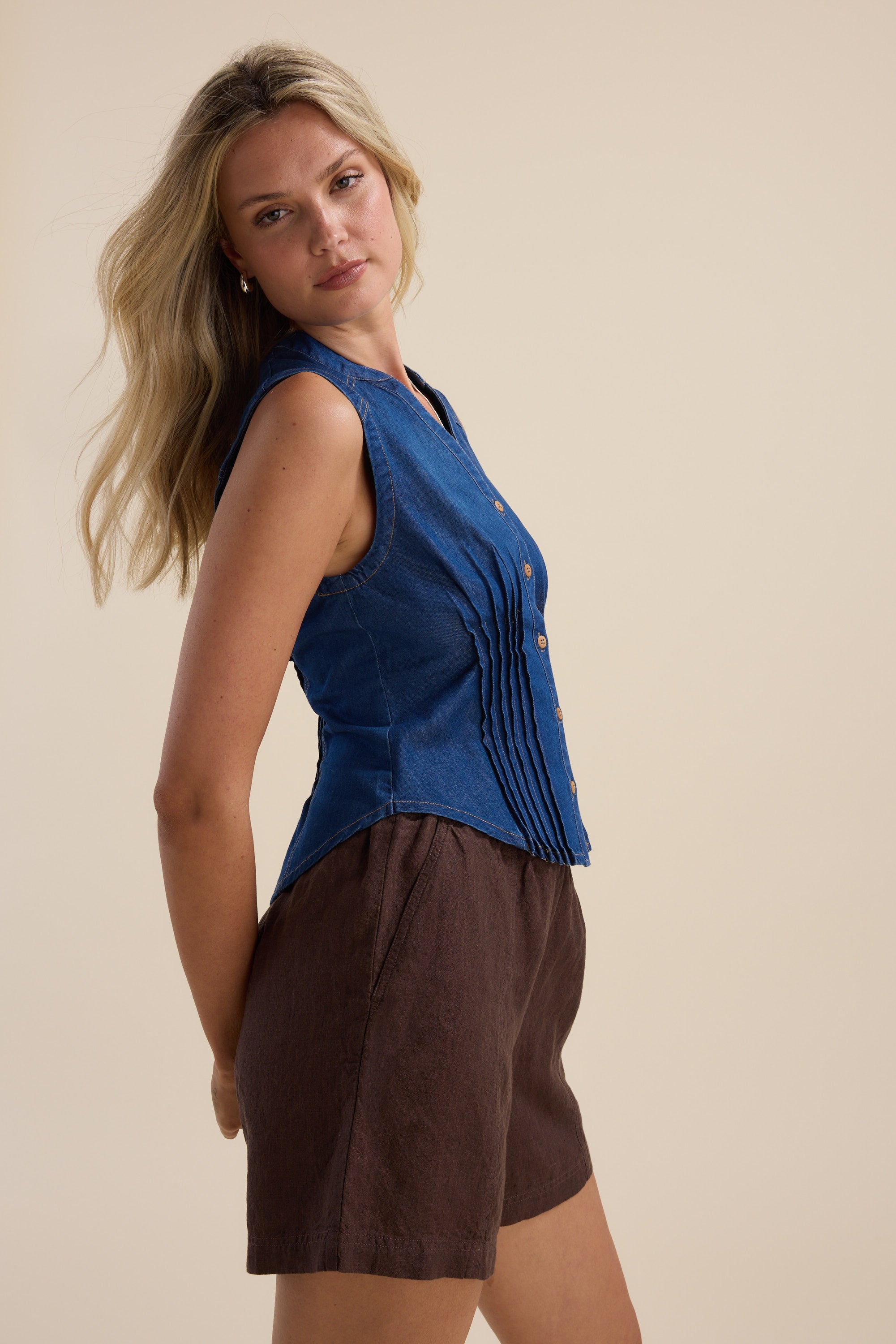 Woman in Brooklyn Tuck Detail Denim Shirt by Max and brown shorts against a beige background.