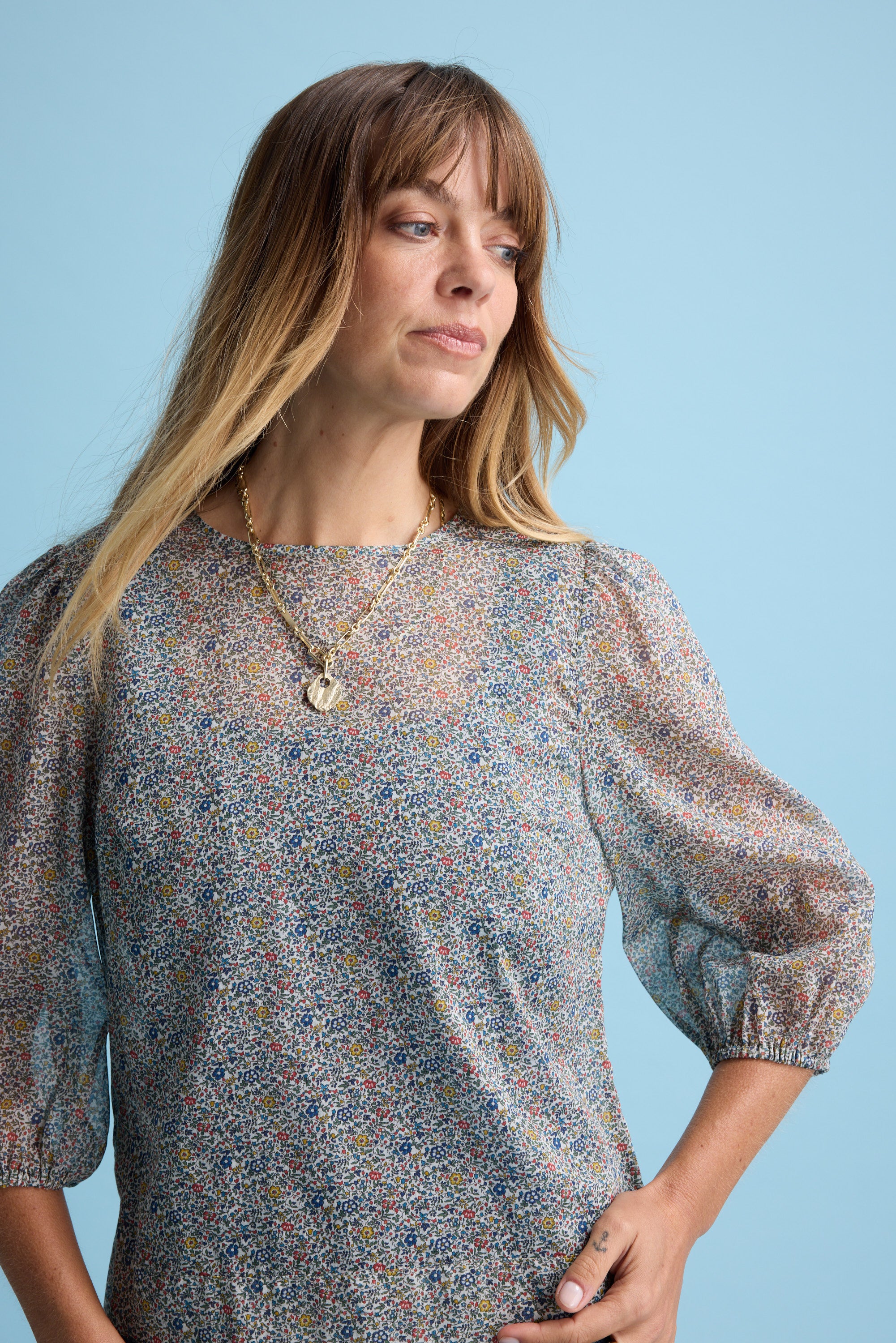 A woman with long, light brown hair wears the Eloise Puff Sleeve Blouse by Max and a gold necklace, standing against a solid light blue background. She gazes slightly to the side with a calm expression.
