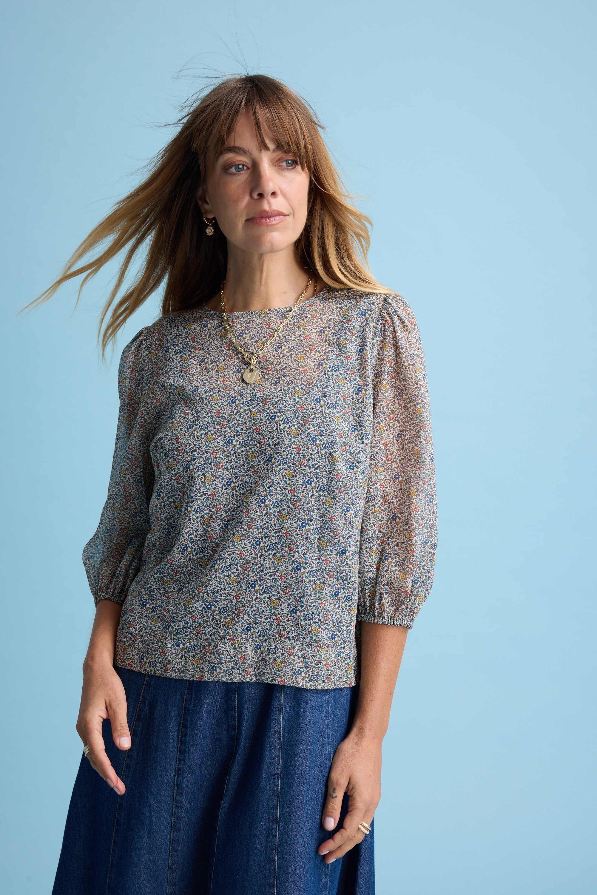 A woman with light brown hair wears the Max Eloise Puff Sleeve Blouse and a blue skirt against a light blue background. Her hair blows gently as she looks to the side, accessorized with layered necklaces and minimal makeup.