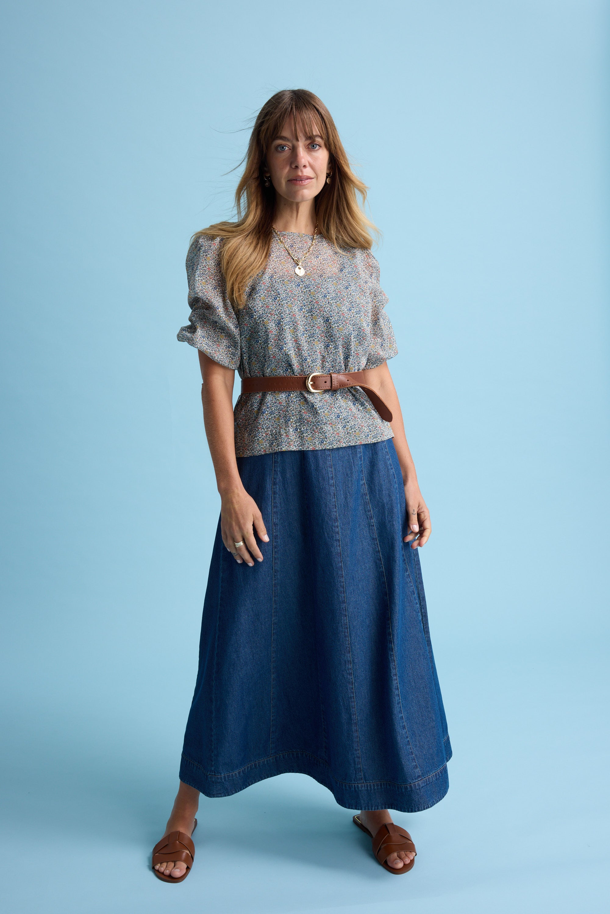A woman stands against a light blue background in the Max Eloise Puff Sleeve Blouse, paired with a brown belt, long denim skirt, brown sandals, and layered necklaces. She looks ahead with a slight smile.