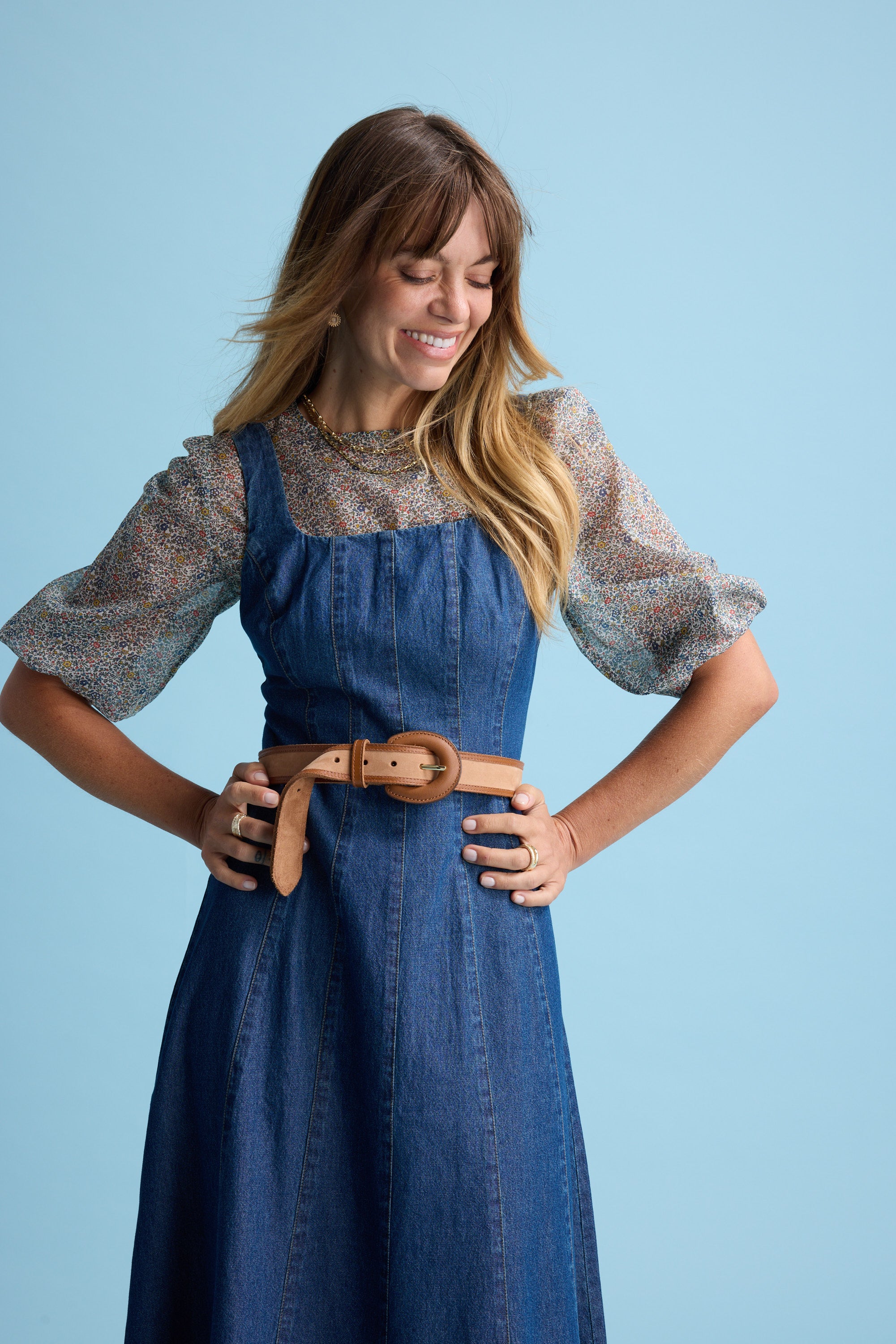 A woman with long light brown hair smiles while looking down, wearing the Max Eloise Puff Sleeve Blouse under a sleeveless denim dress with a tan belt, standing against a light blue background.