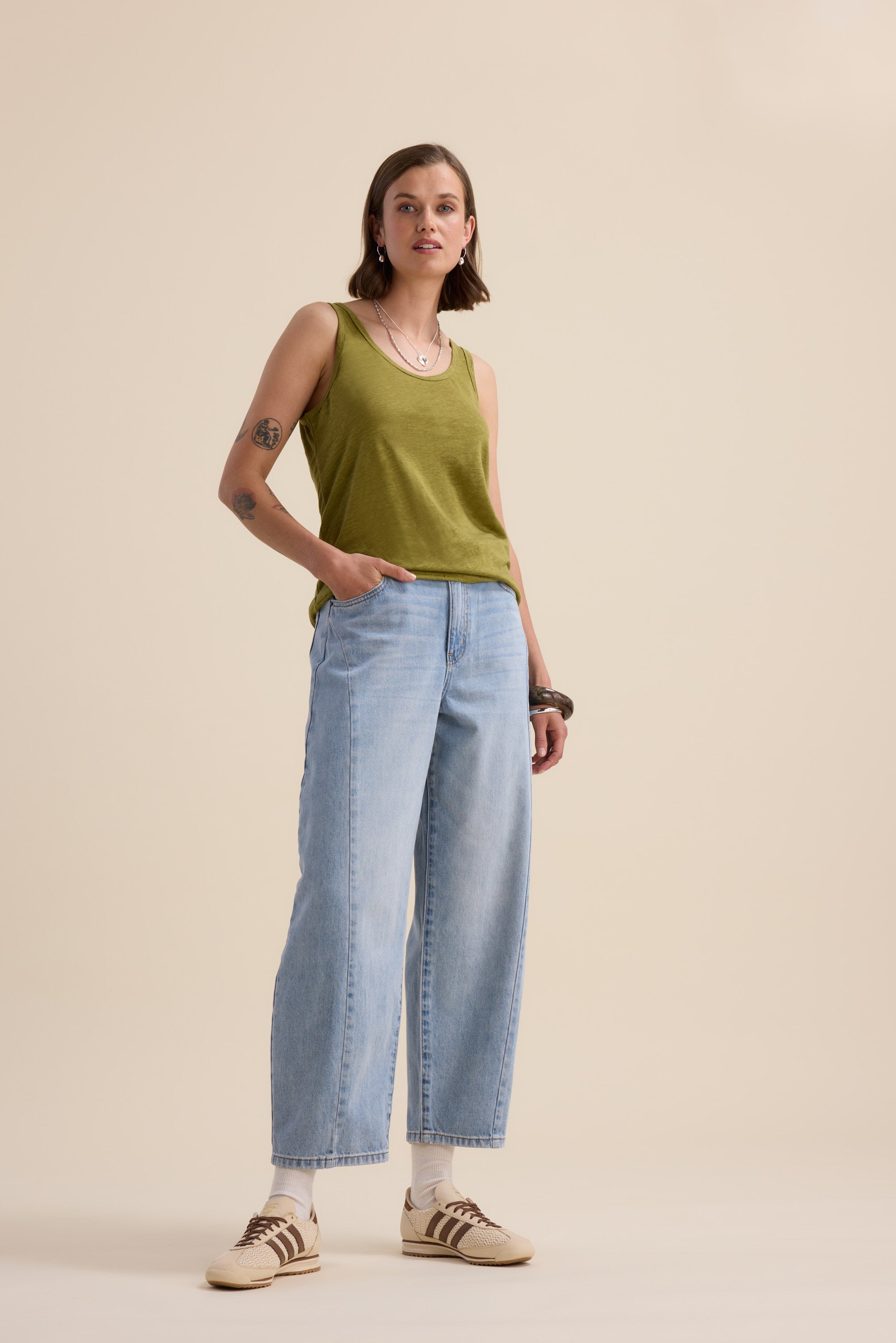 A person with short hair, tattoos, and jewelry stands against a beige background wearing the Elementary Linen Jersey Tank by Elementary, styled with light blue wide-leg jeans, white socks, and beige sneakers—one hand in their pocket.