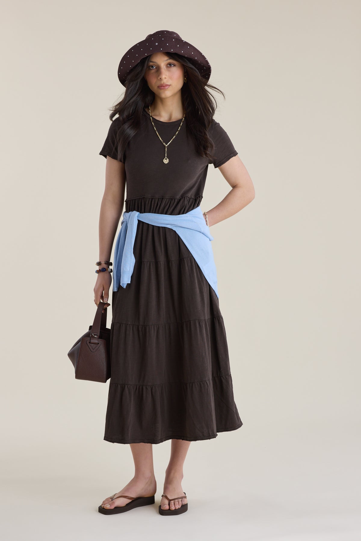Woman in an Elementary Jersey Tiered Dress, blue sweater at waist, brown hat, brown bag.