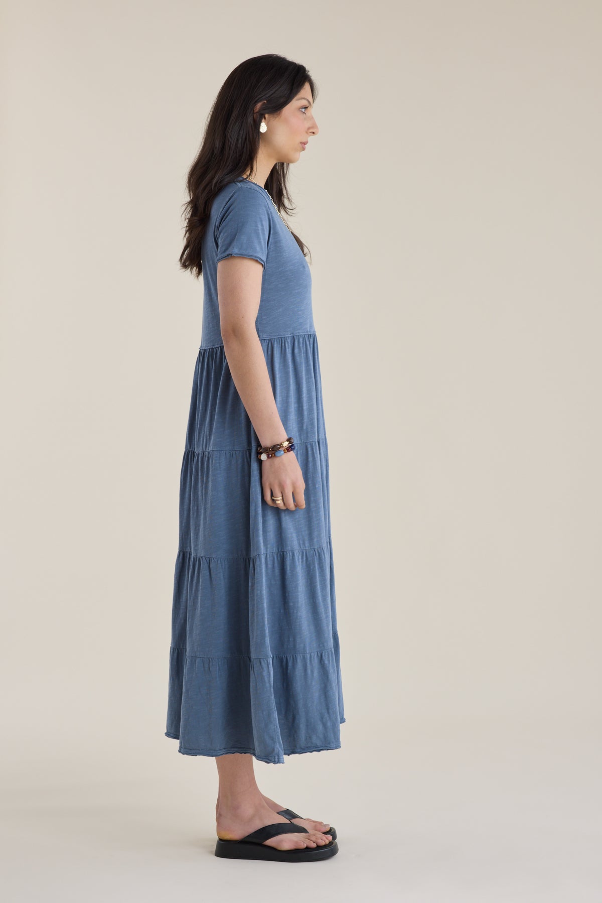 Woman in an Elementary Jersey Tiered Dress and black sandals stands in profile on beige.