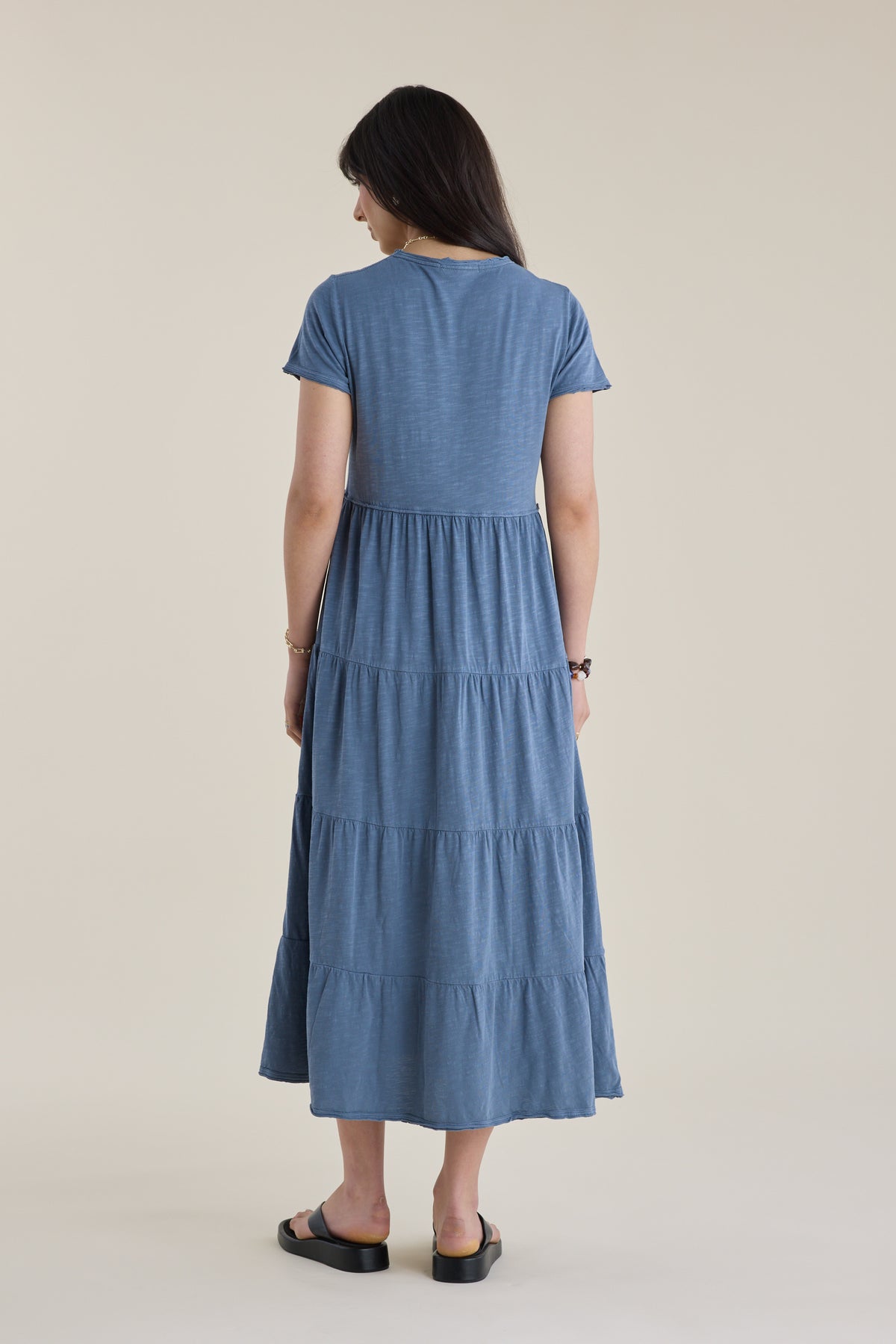 Woman in an Elementary Jersey Tiered Dress stands facing away on a plain background.