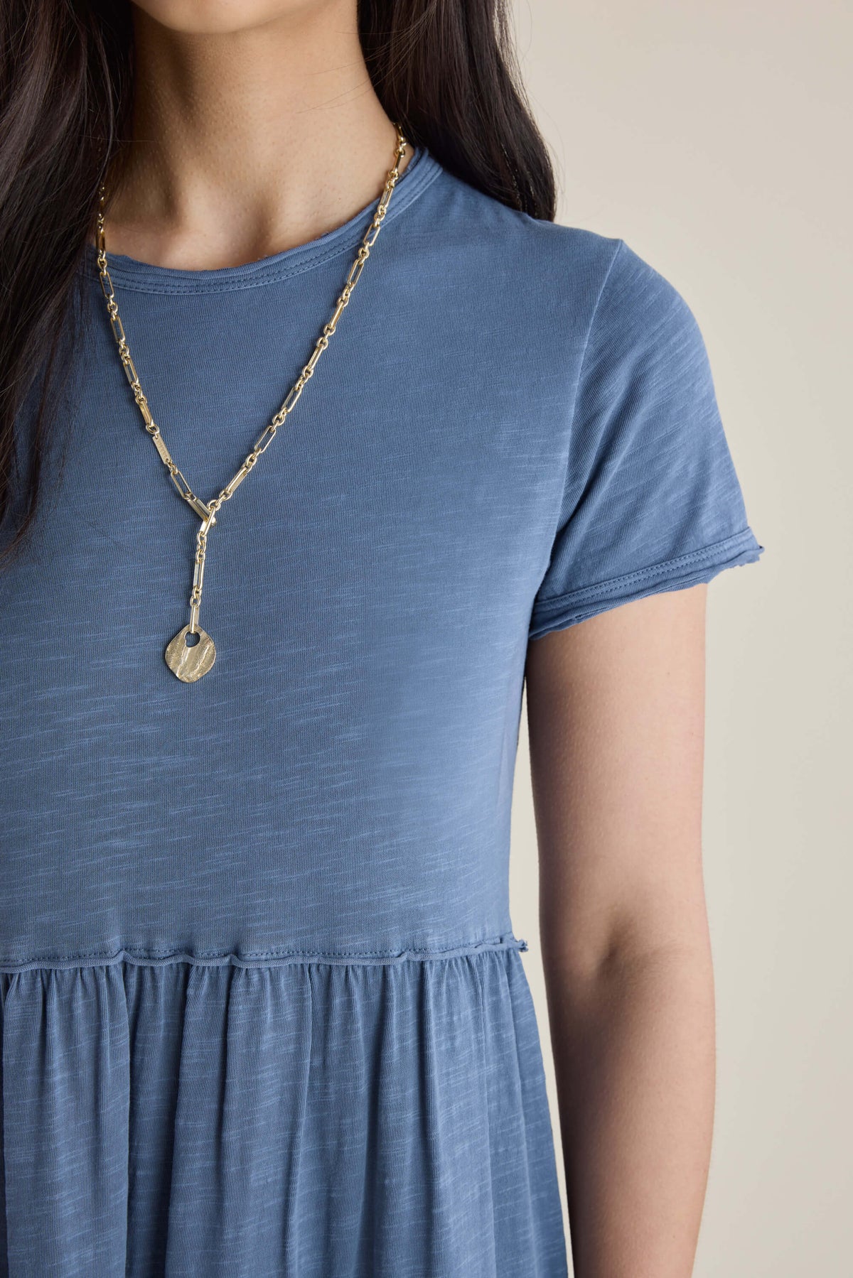 Woman in an Elementary blue jersey tiered dress, paired with a gold heart pendant necklace.