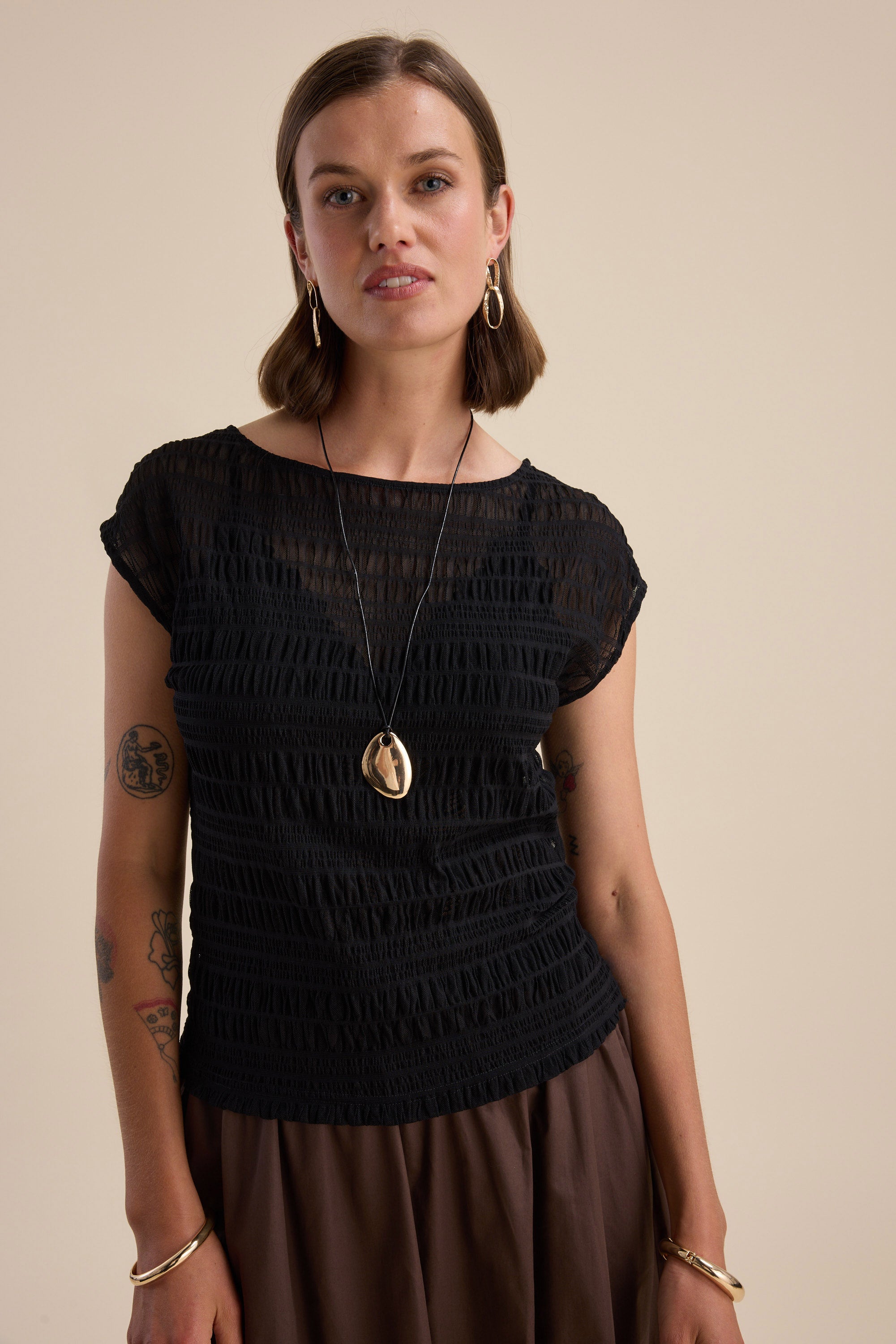Woman in Max's Willow Textured Mesh Tee and brown skirt, gold jewelry, beige backdrop.