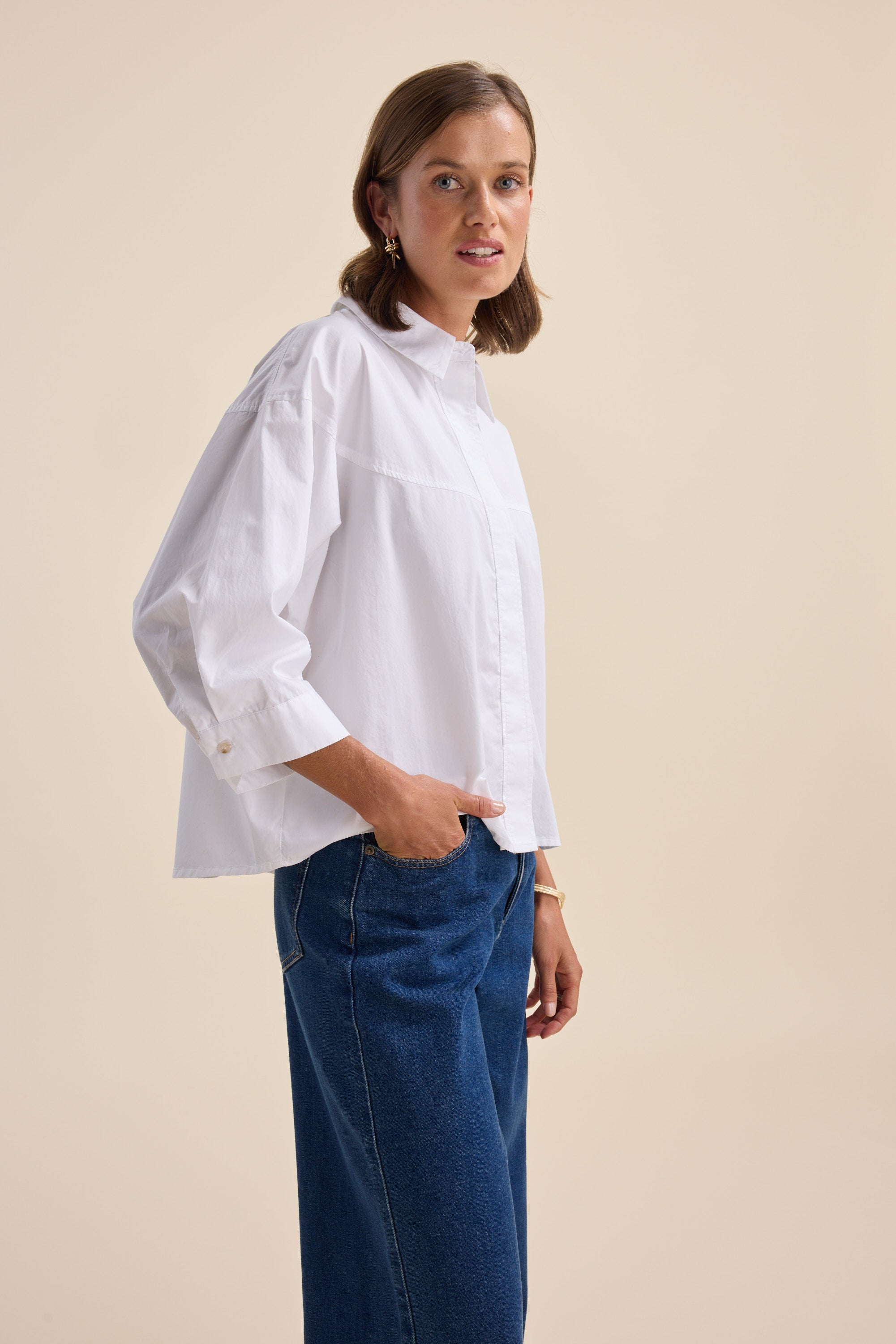 Woman in Elementary Yoke Detail Shirt and blue jeans stands against a beige background.