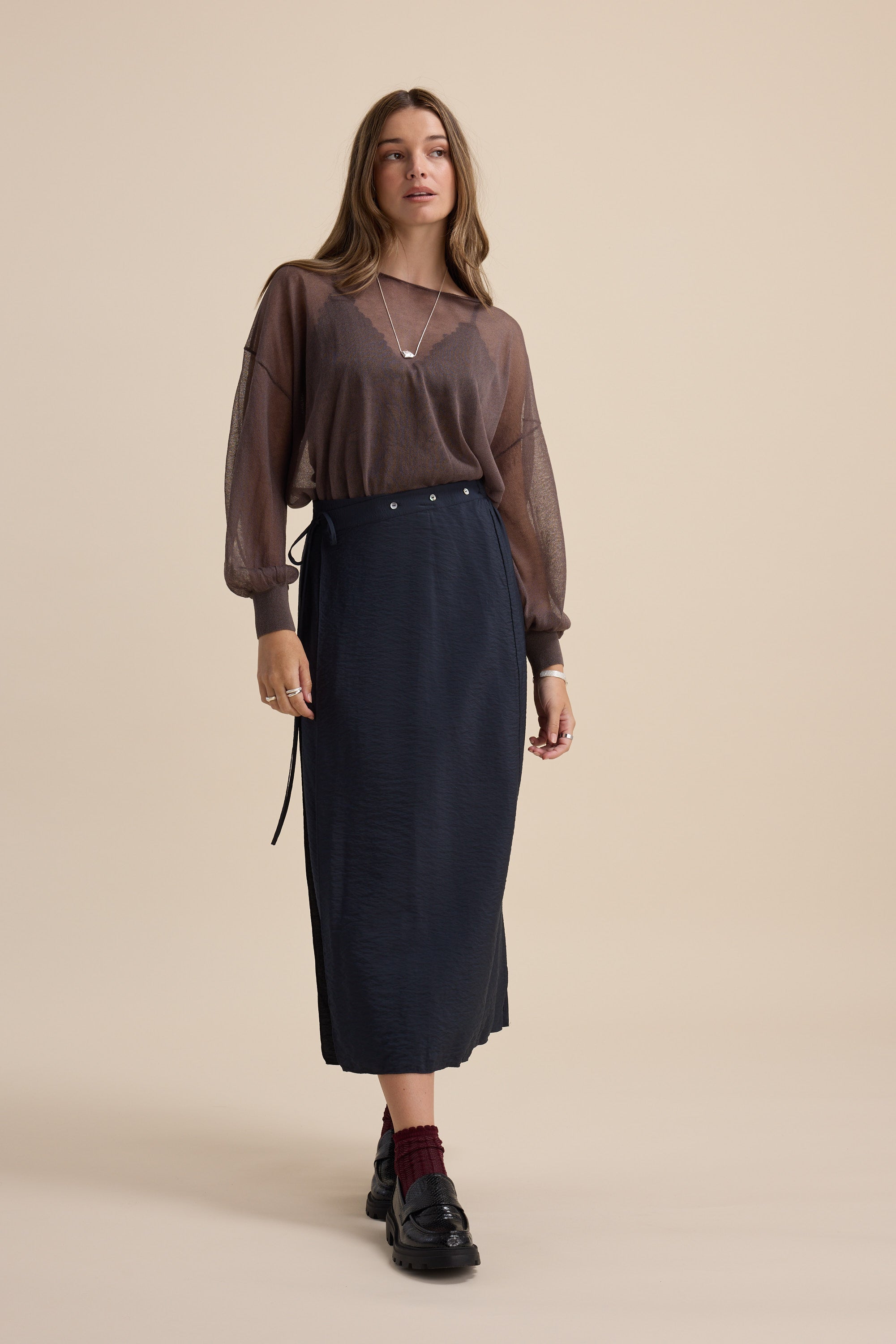 Woman in sheer brown top and Elementary Wrap Detail Skirt by Elementary, against beige background.