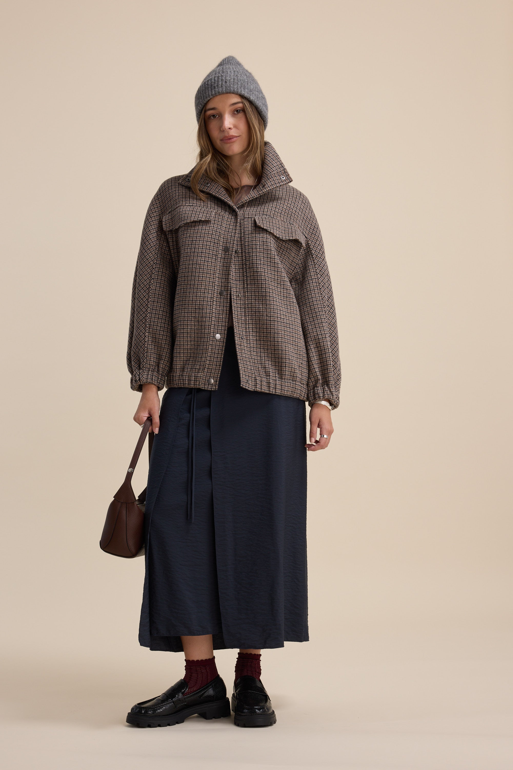 Woman in a beanie, plaid jacket, Elementary Wrap Detail Skirt by Elementary, holding a brown bag.