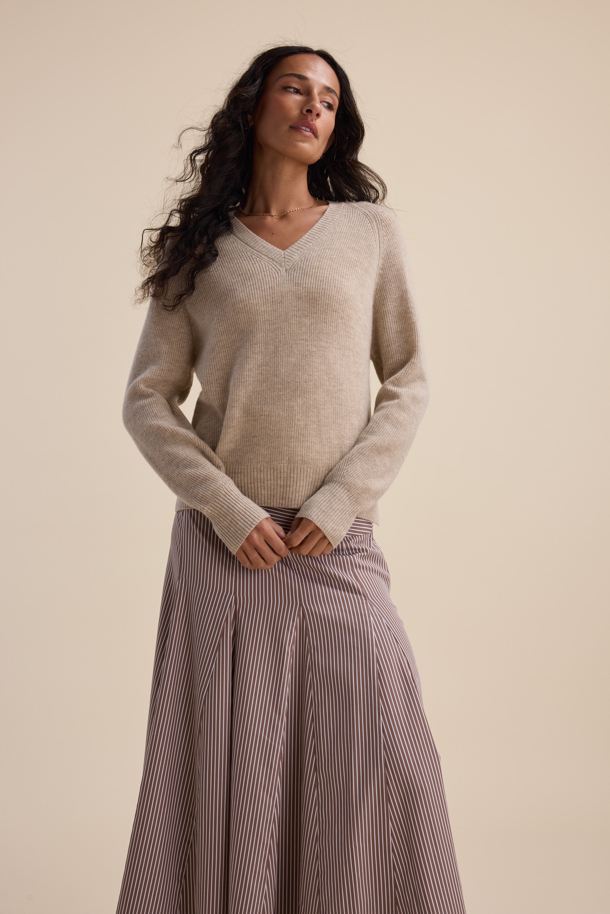Woman wears Elementary Cashmere Blend V Neck Sweater and striped skirt against a beige background.