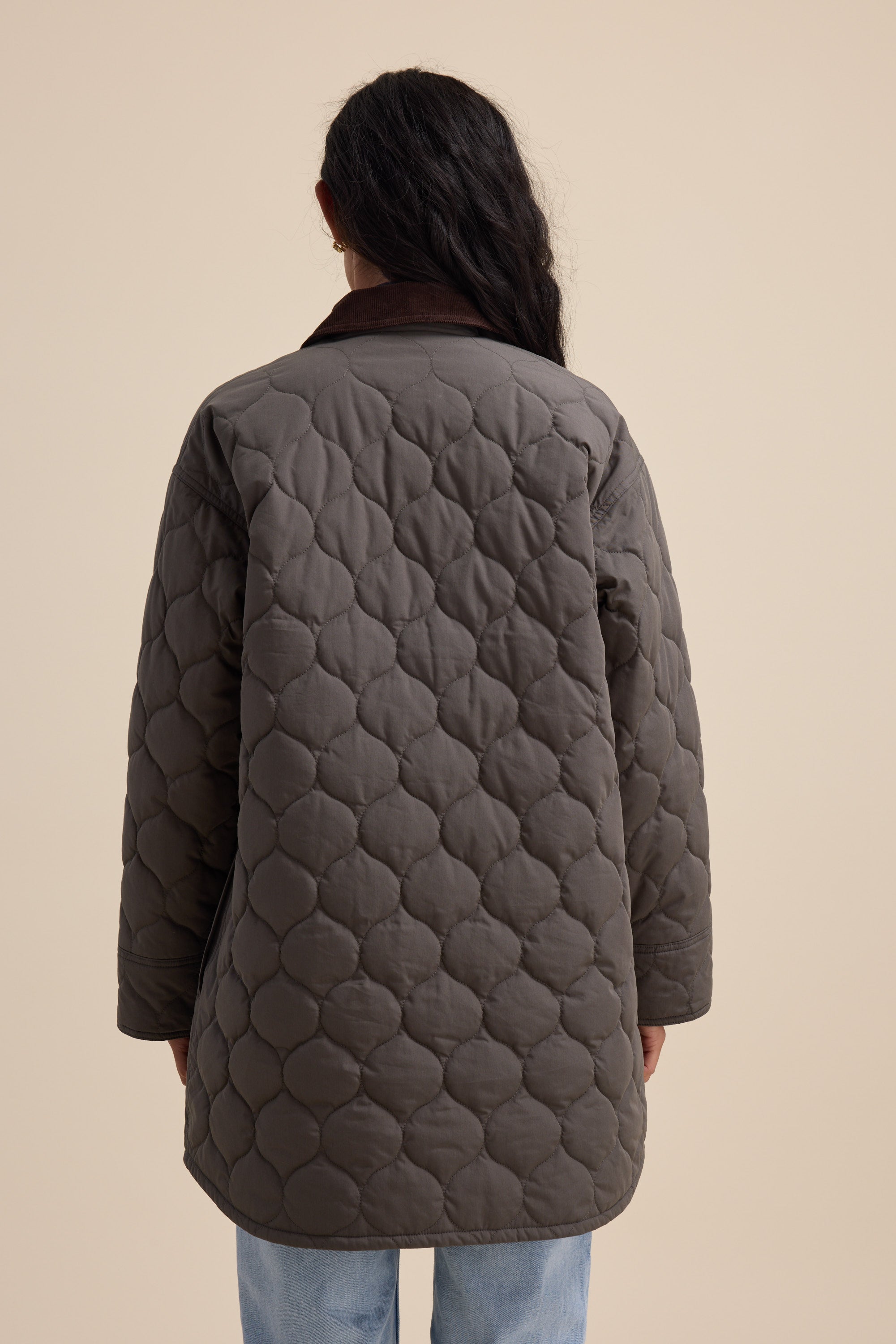 Someone wears the Elementary Longline Puffer in gray, shown from the back on a beige background.
