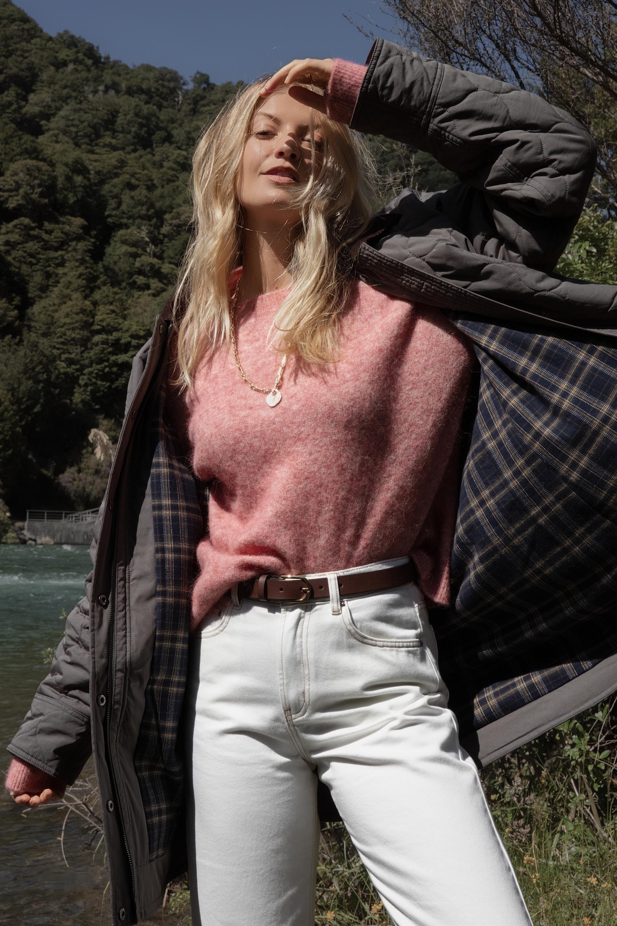 Woman in Elementary's Longline Puffer, pink sweater, and white pants stands outdoors.