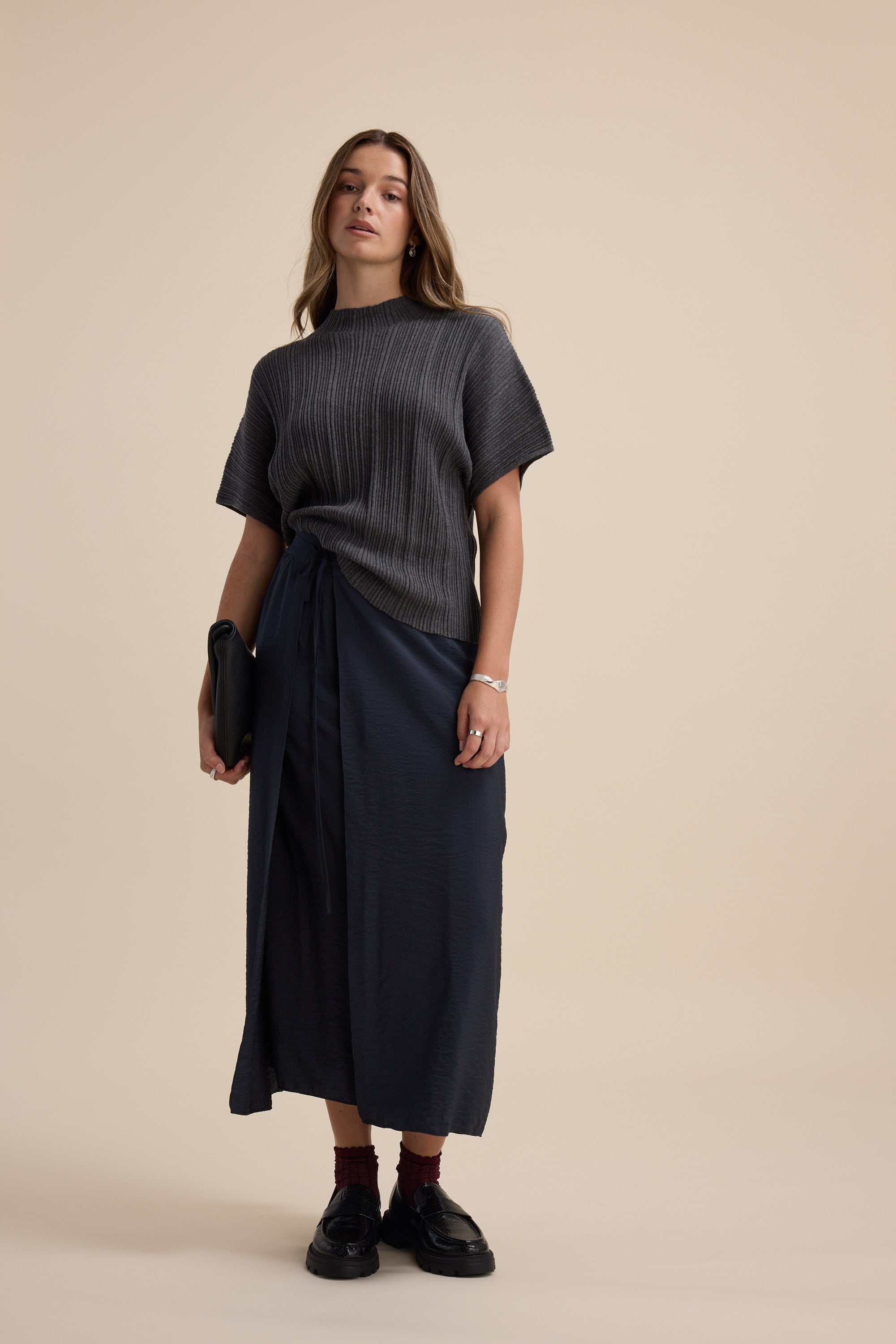 Woman in gray knit top and Elementary Wrap Detail Skirt, holding a black clutch on beige.