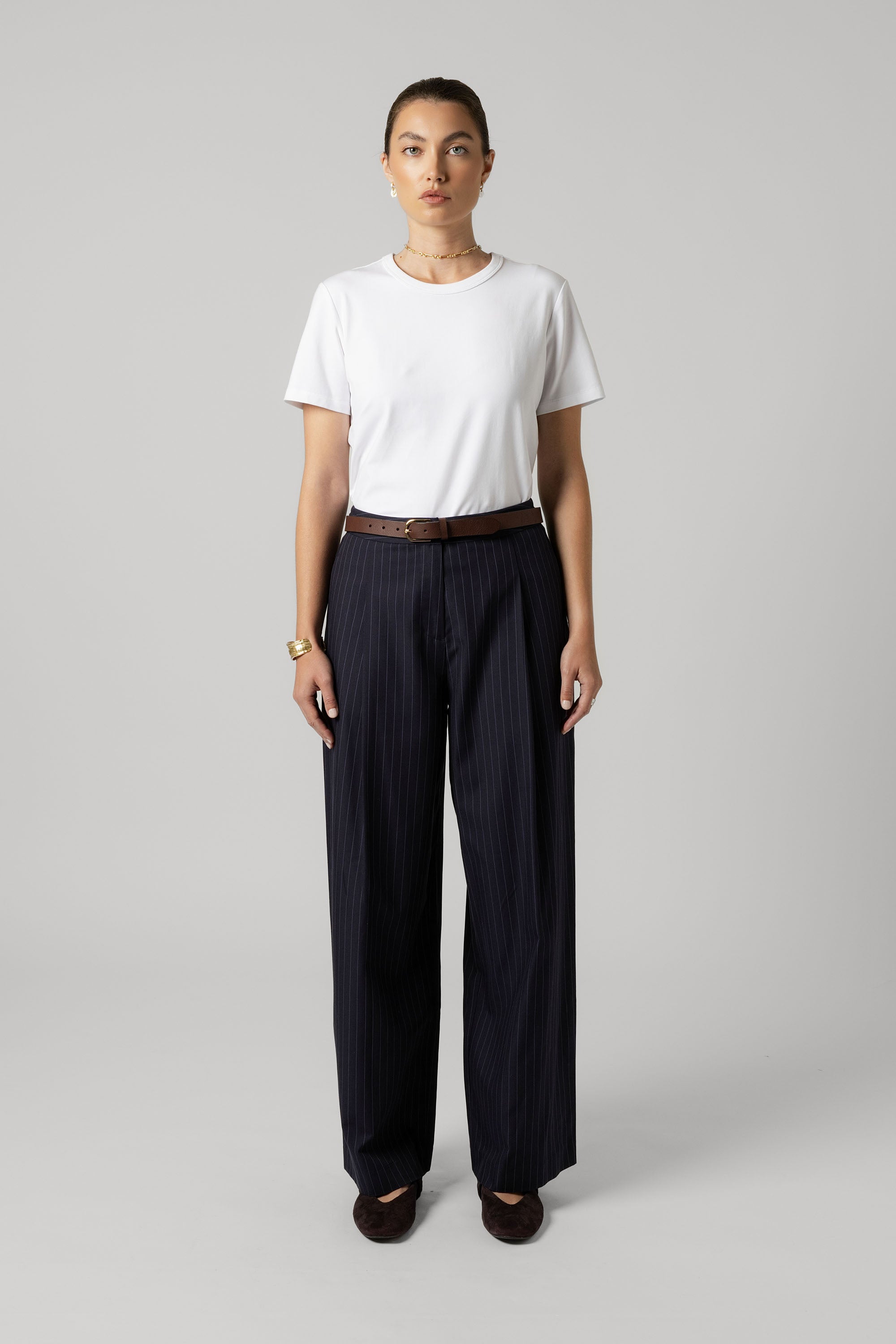 Woman in white tee and Max The Pleat Detail Wide Leg Pants with brown belt, gray background.