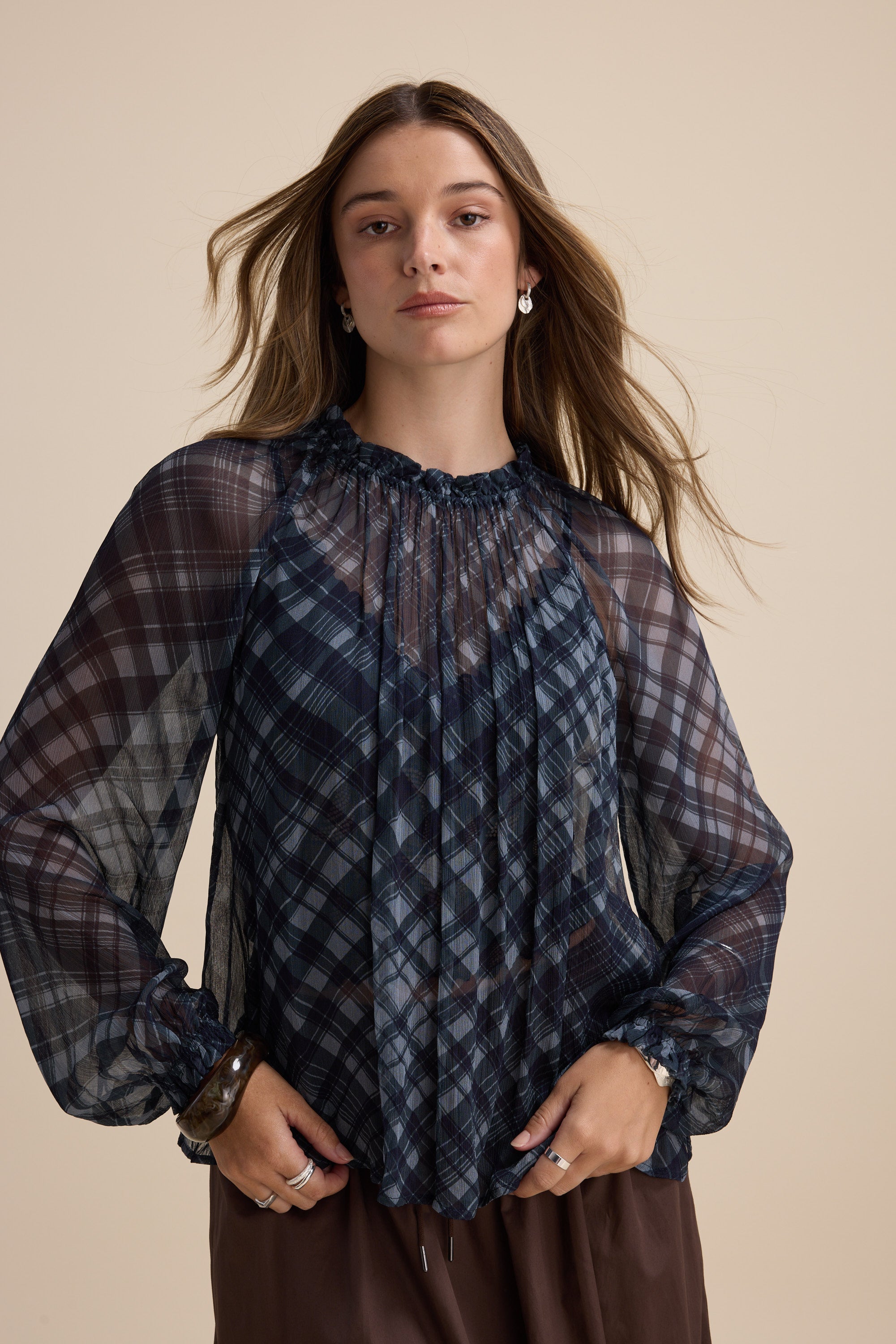 Woman in Max's Quinn Gather Detail Blouse, navy check with sheer sleeves, on beige background.