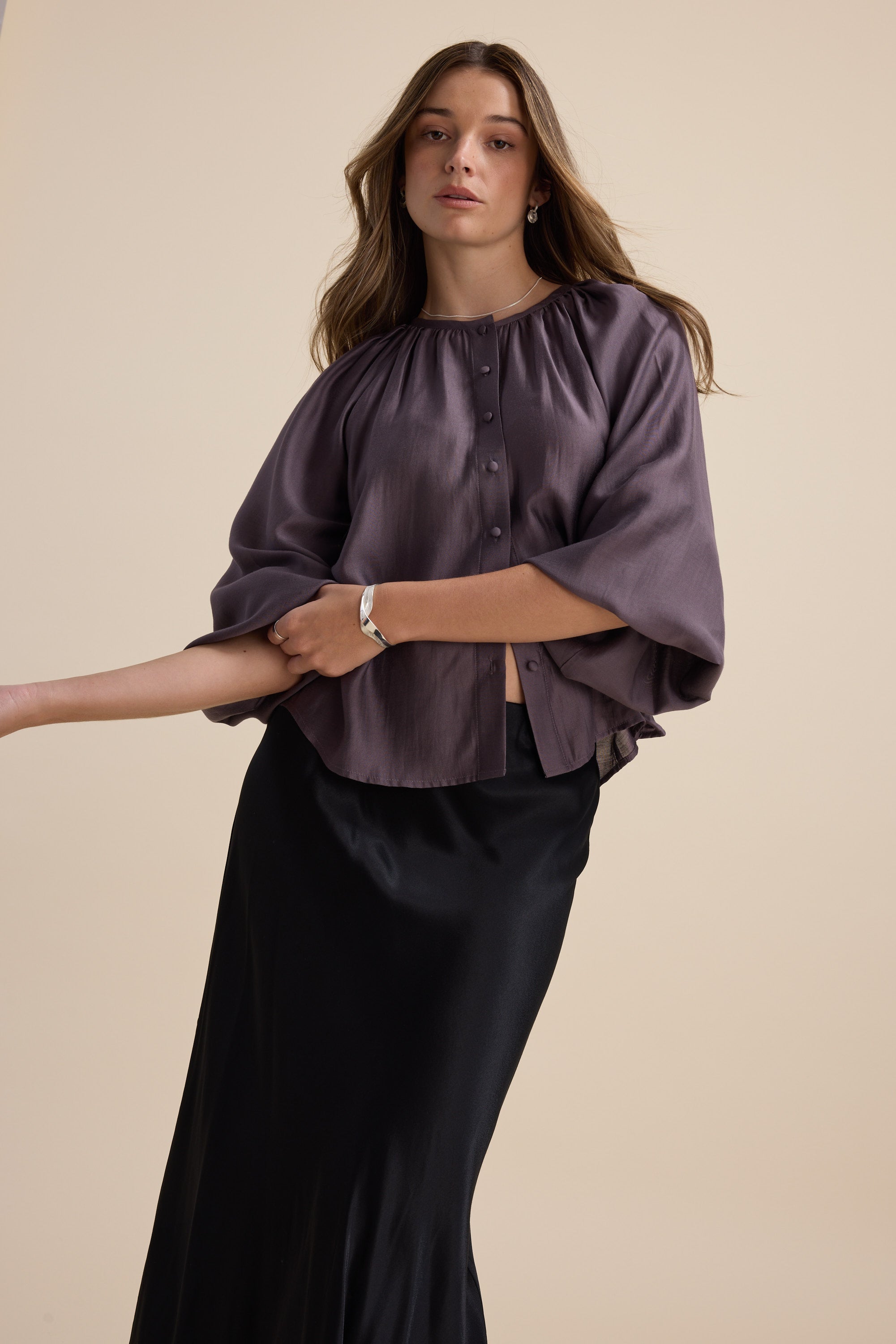 Woman in a loose Vivian Button Front Blouse by Max and black skirt poses on beige background.