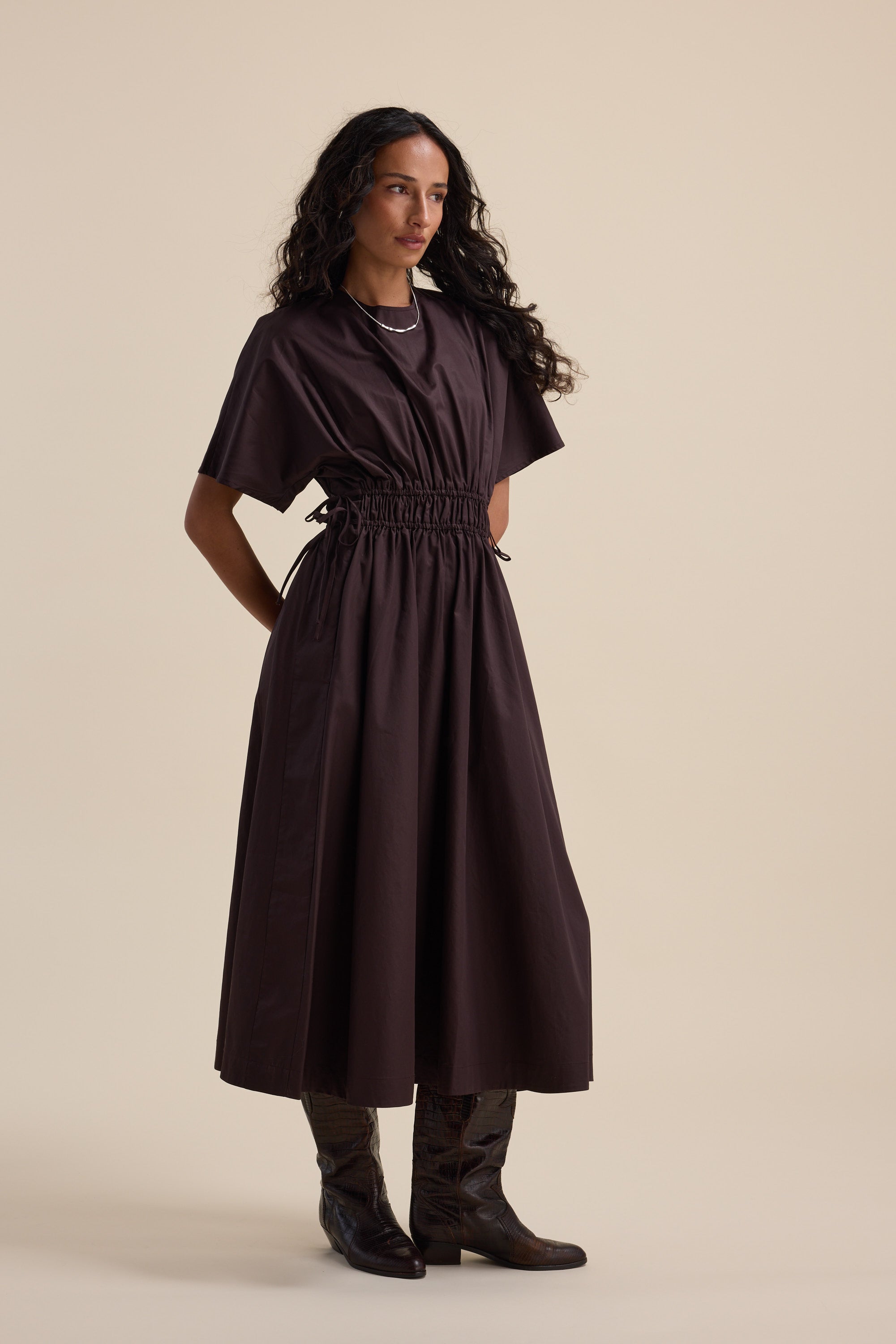 Woman wears the Max Megan Tie Waist Dress, styled with boots against a beige background.
