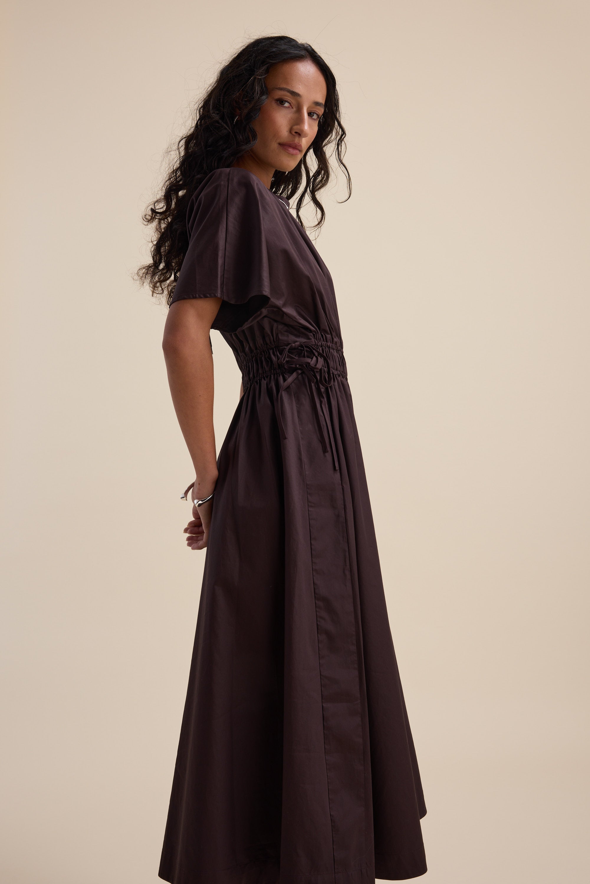 Woman wears Max Megan Tie Waist Dress in organic cotton, dark brown, against beige background.