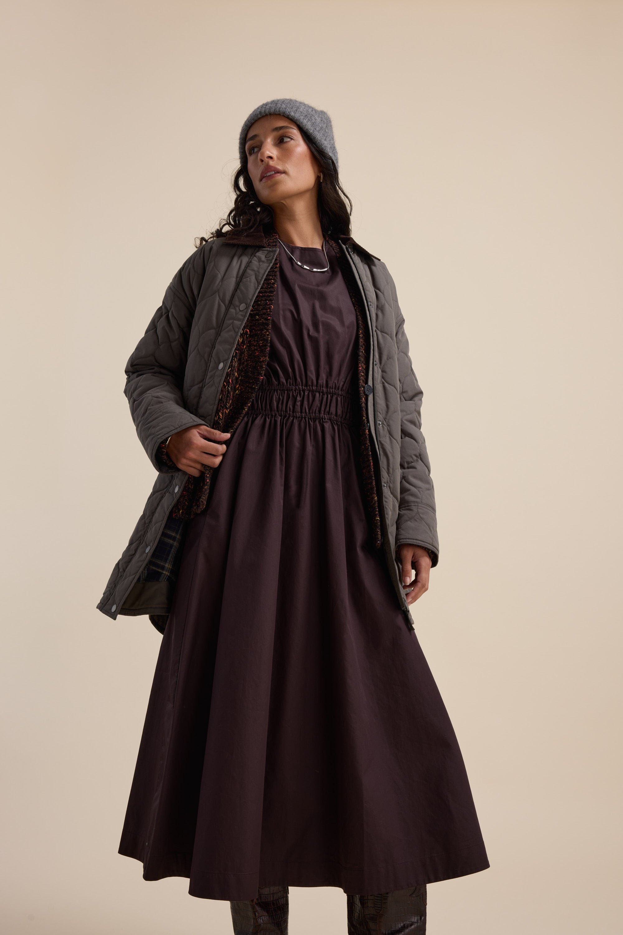 Woman in a Max Megan Tie Waist Dress, gray quilted jacket, and beanie stands confidently.