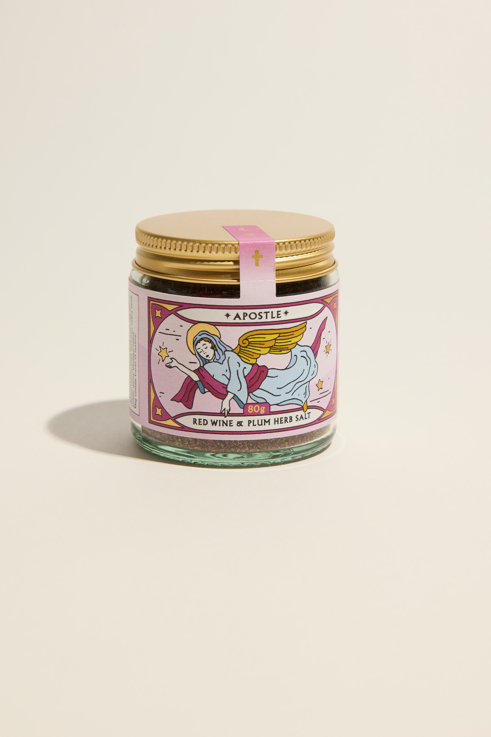 A glass jar with a gold lid displays a pink label featuring an angel and gold accents. The label reads "Apostle Red Wine & Plum Herb Salt" by Apostle, crafted with Provençal herbs. The jar is set against a plain, light background.