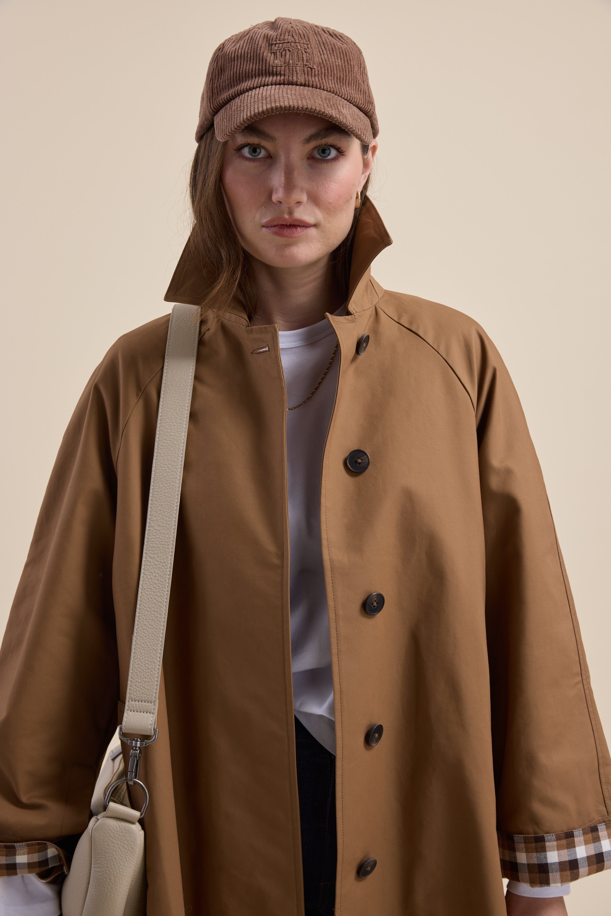 Woman in a mid length London Trench Coat by Max, beige bag in hand, faces the camera.