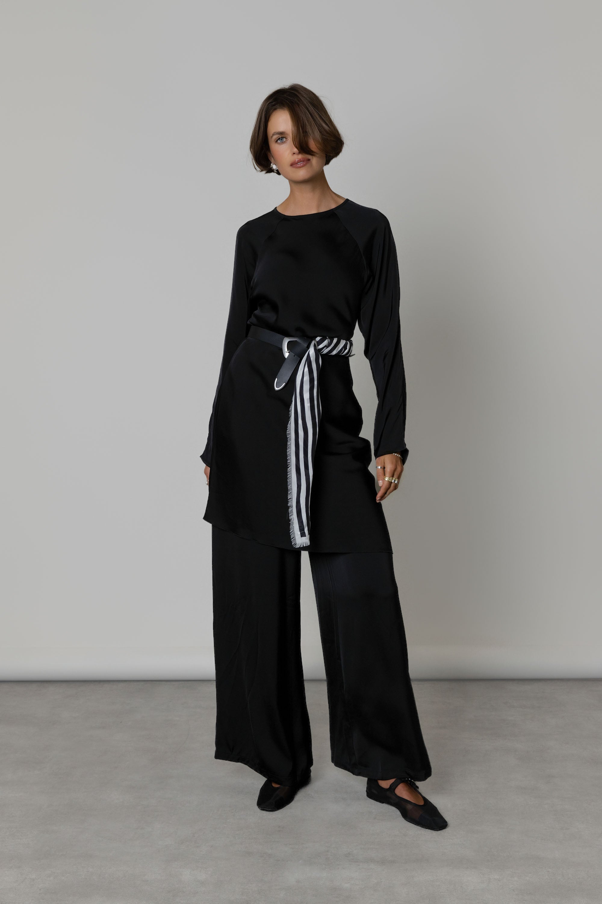 Woman in black outfit with wide pants, striped belt, and Max Mable Frayed Edge Silk Scarf indoors.