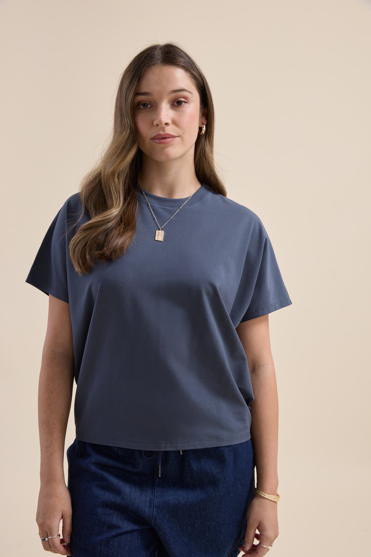 Woman wears Elementary Slouchy Short Sleeve Tee and jeans, standing against a beige background.