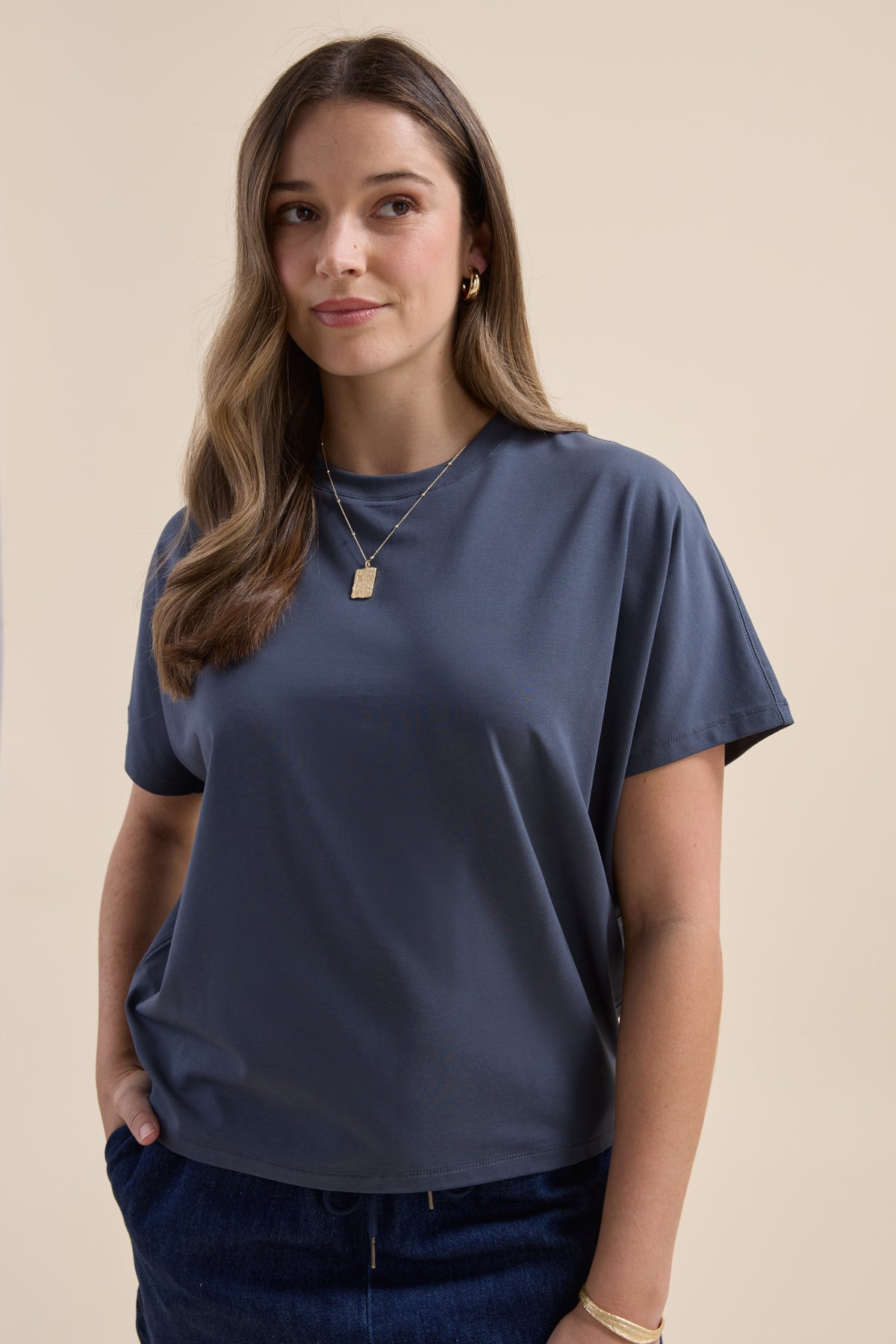 Woman wears Elementary Slouchy Short Sleeve Tee and dark pants, hands in pockets, neutral backdrop.