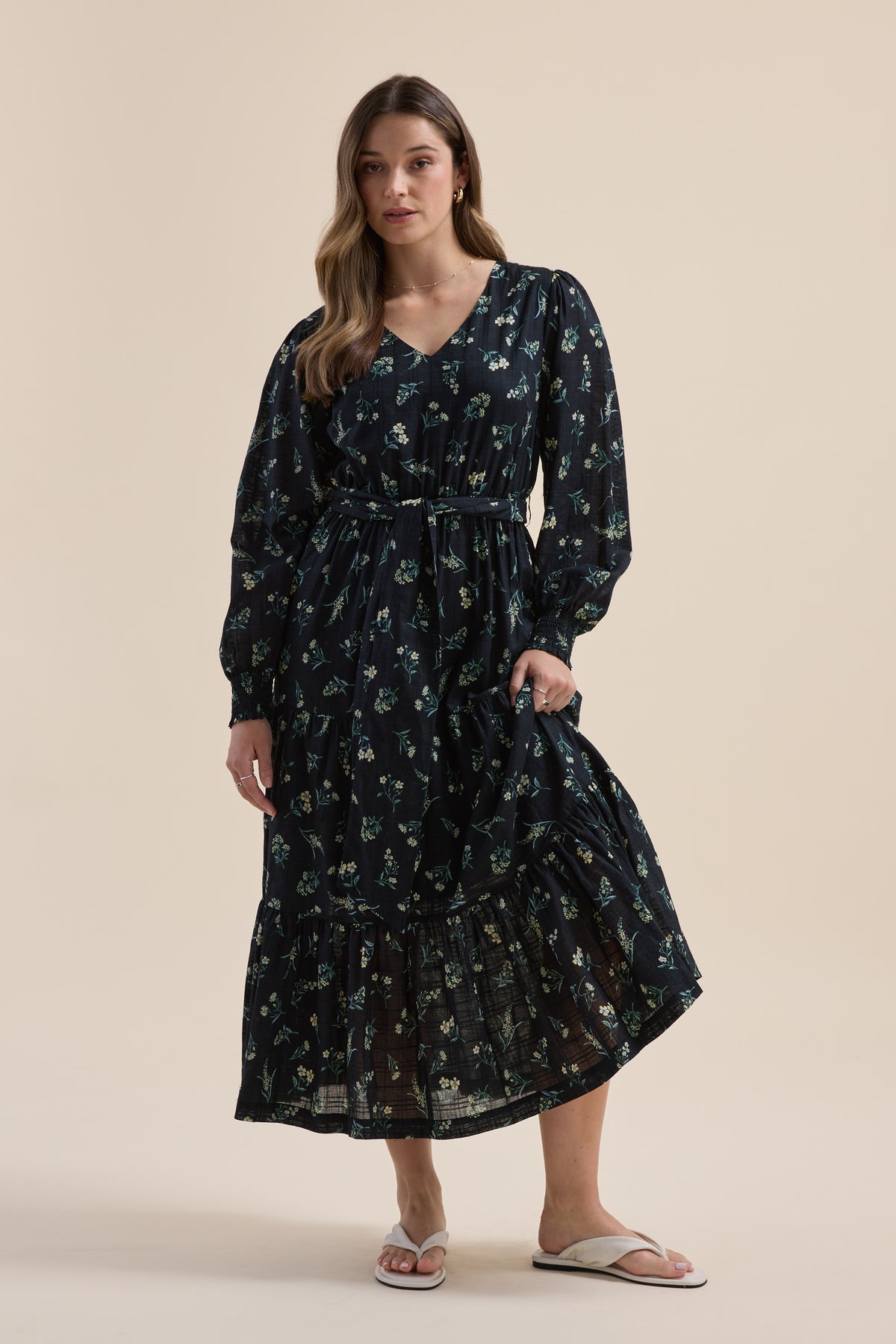 Woman wears Elementary Tiered Dress with daisy print and sandals against a plain background.