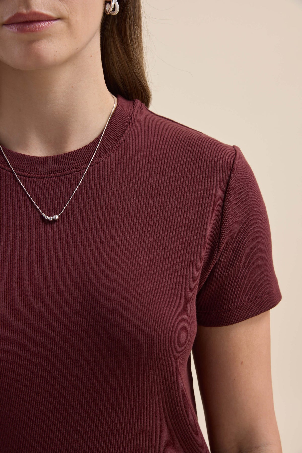 Woman in maroon Elementary Ribbed Boxy Tee, neck to mid-arm, beige background, wearing necklace.
