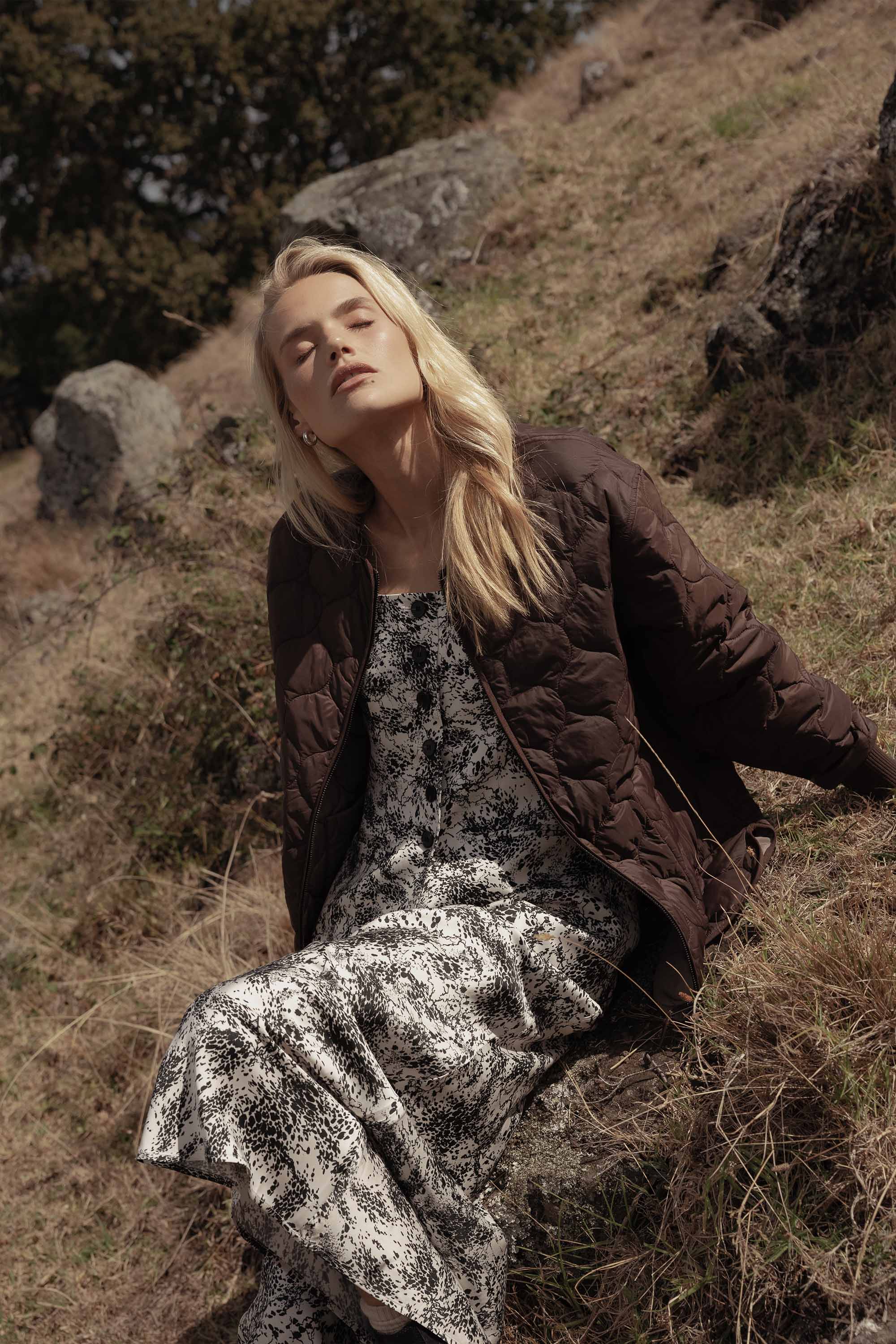 Woman in Max’s Henry Maxi Slip Skirt and brown jacket sits on grass, eyes closed in sunlight.