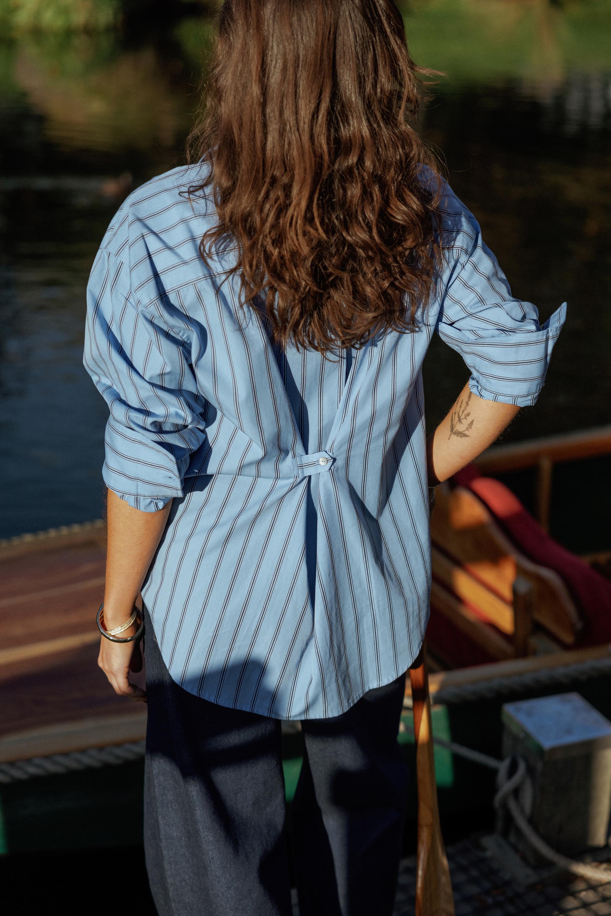 Person with wavy brown hair wears the Max Cameron Oversized Shirt by a docked wooden boat.