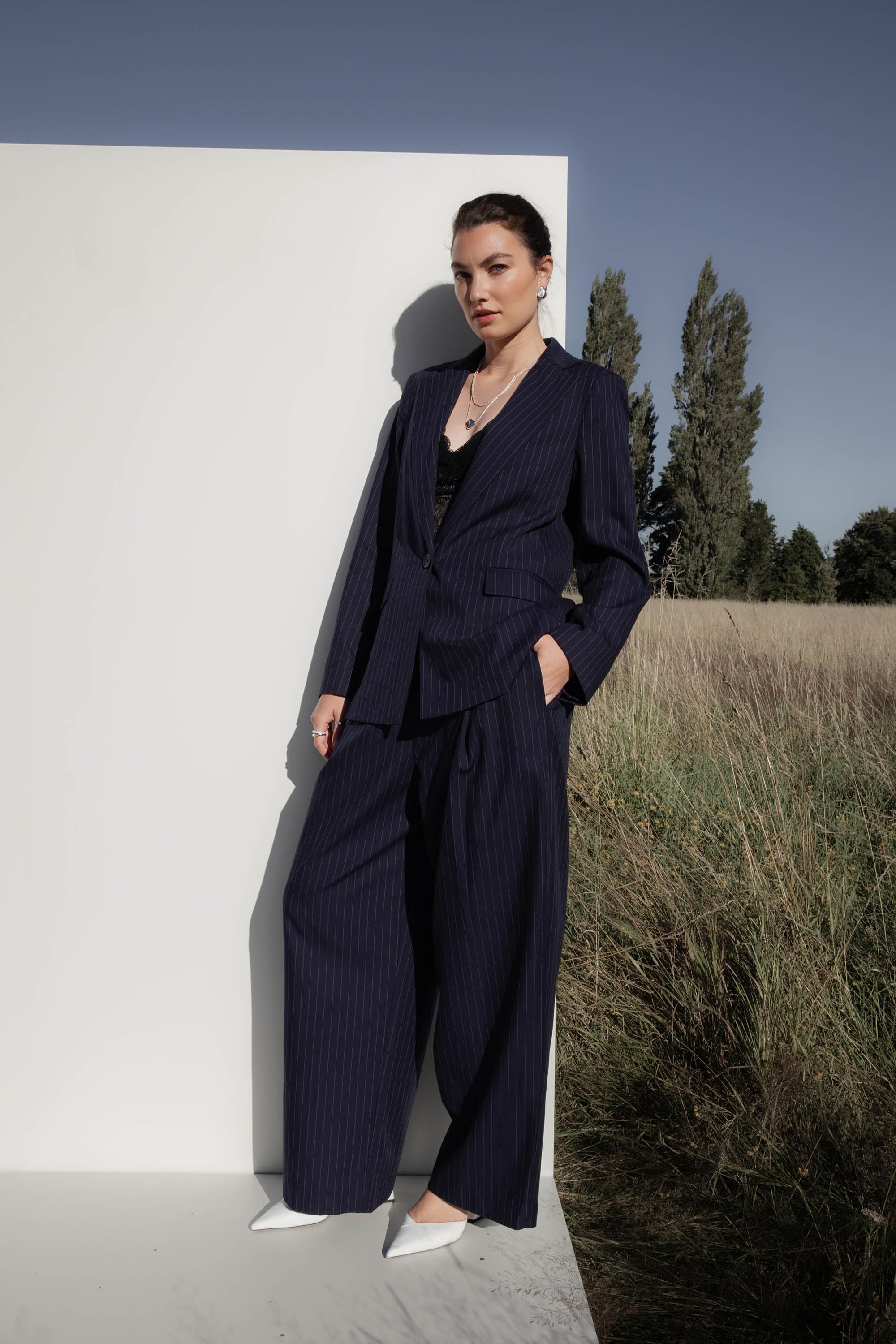 Woman wears Max’s Pleat Detail Wide Leg Pants in navy pinstripe, standing by a white wall.