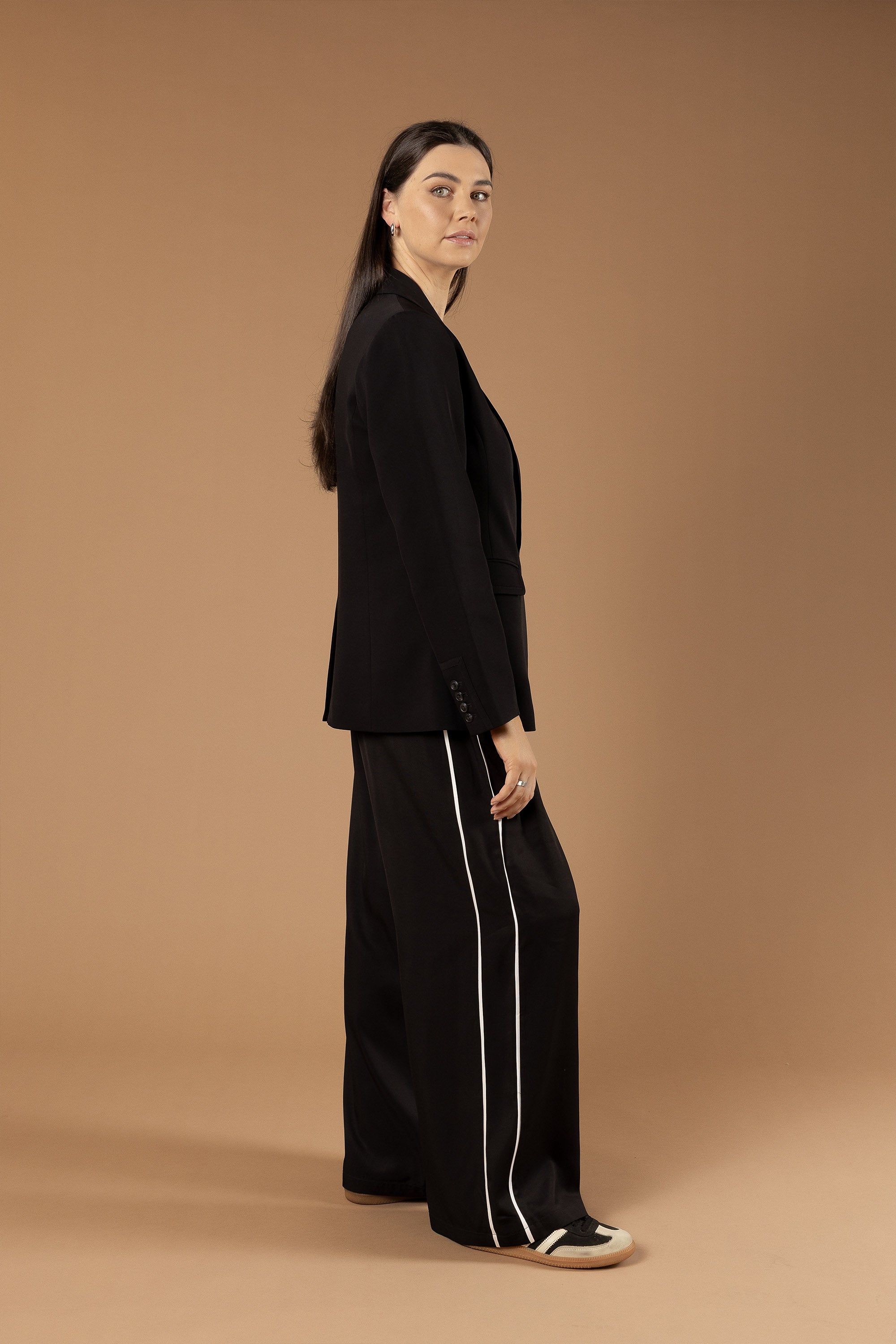 A woman with long brown hair stands sideways against a beige background, wearing Max's The Elevated Jogger, a high-rise wide leg pant with white stripes, paired with a black blazer and black sneakers with white soles.