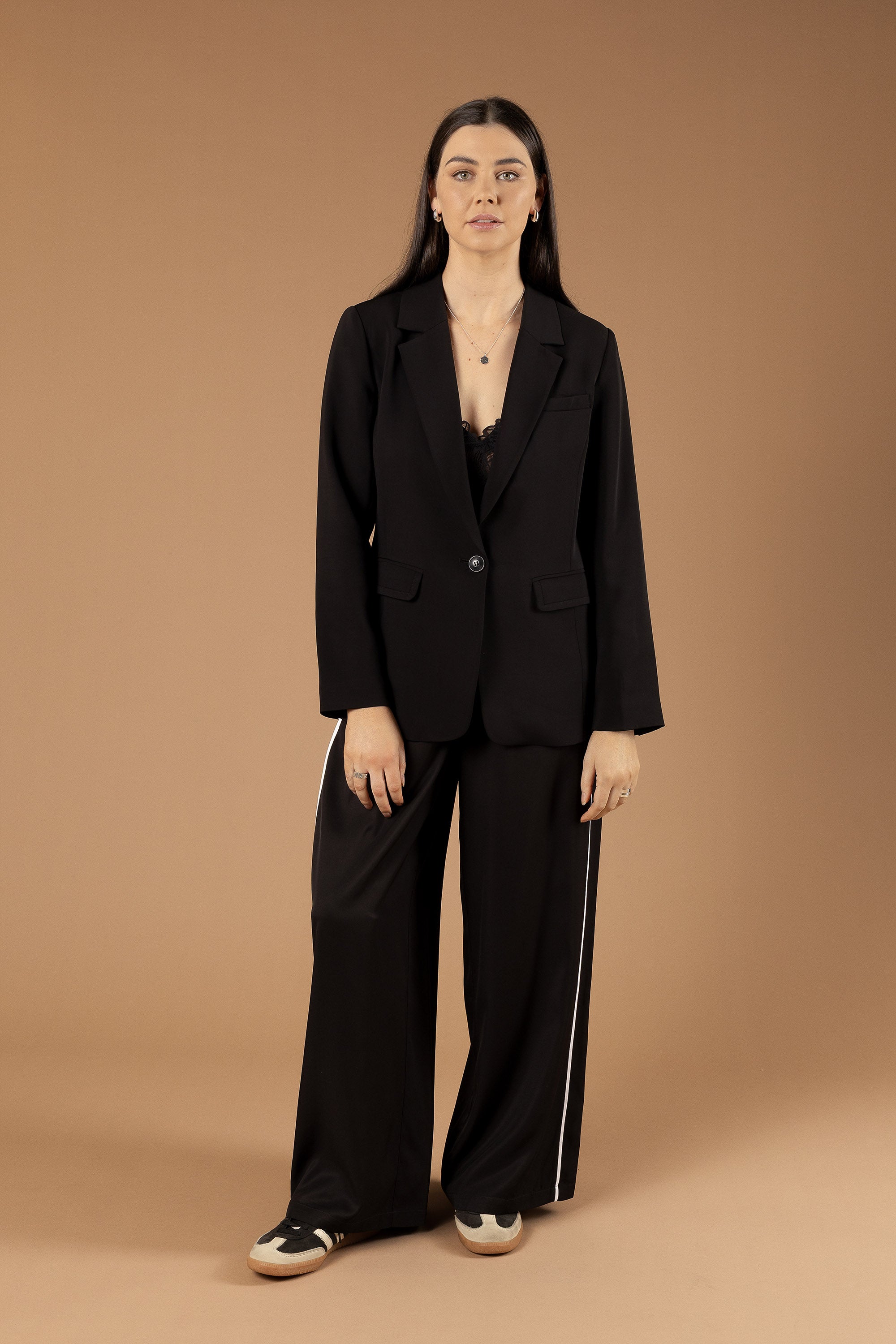 A woman with long straight hair and a neutral expression wears Max’s The Elevated Jogger—black jogger pants with white stripes—paired with a black blazer, lace top, and black-and-white sneakers against a plain brown background.