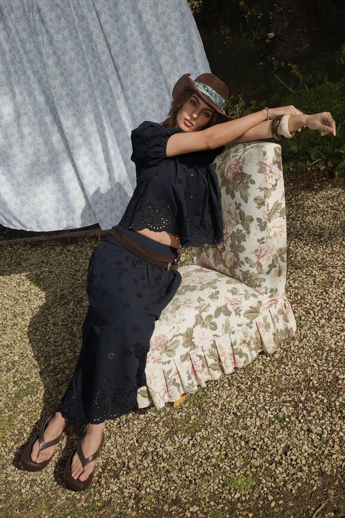 Woman wears Elementary Broderie Detail Skirt, lounging outdoors on floral chair by hanging sheet.