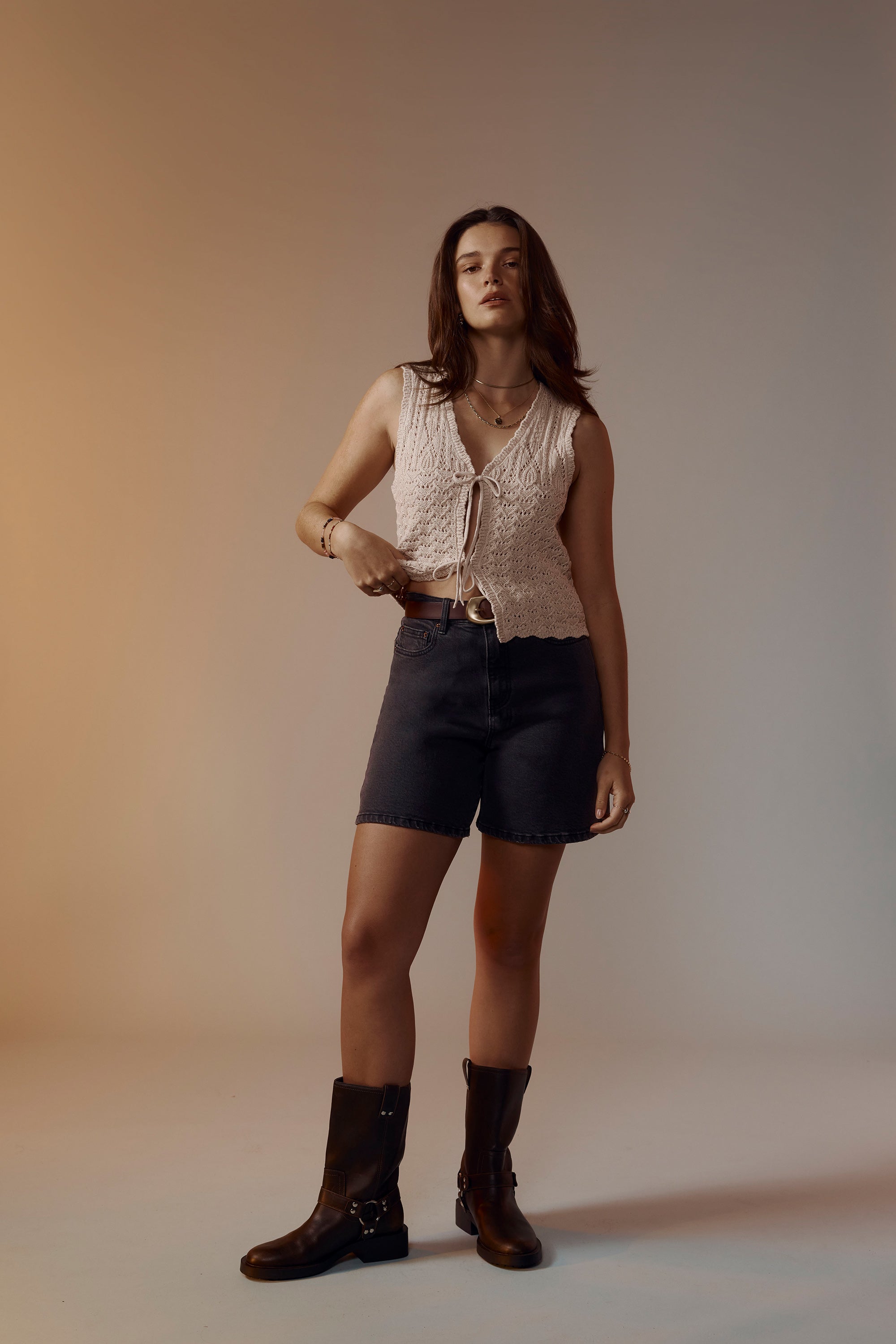 A woman stands in a studio wearing Max's The Ultimate Denim Short in dark denim with a sleeveless top and black boots. She has medium-length brown hair and looks at the camera with a neutral expression.