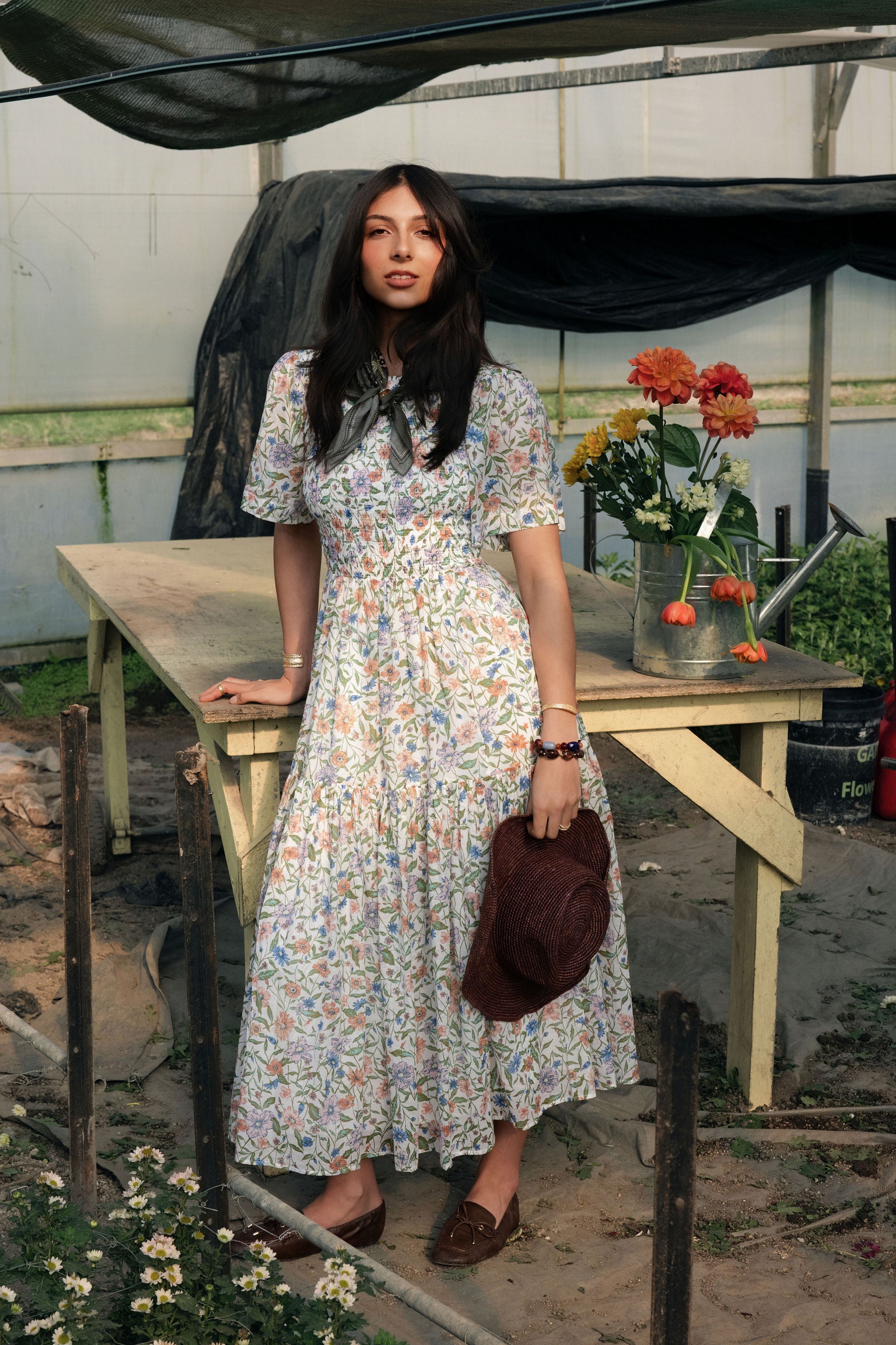 A woman standing in a greenhouse wearing a floral spring midi dress and a green scarf.